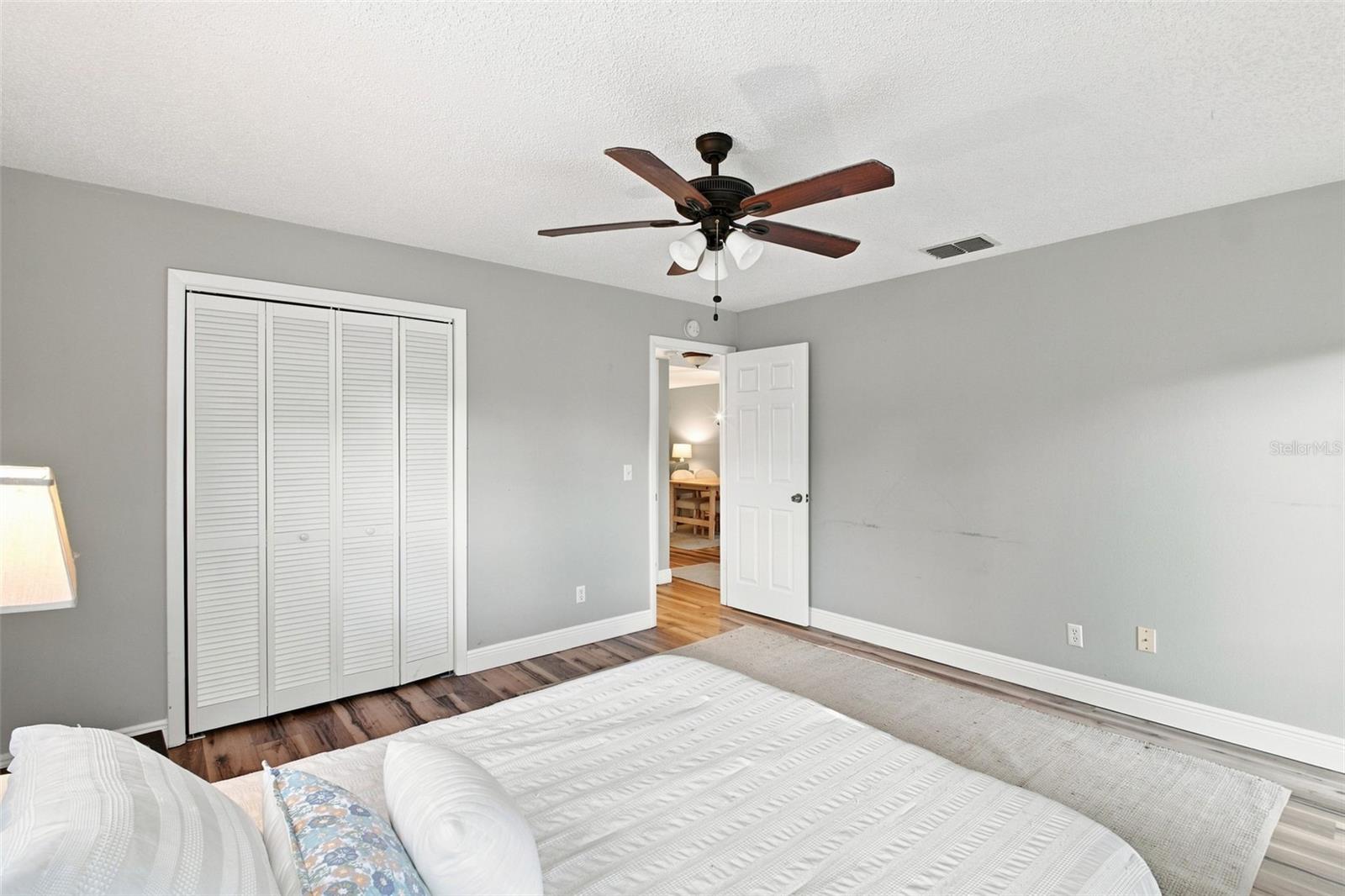 Furnished bedroom with a white bed, ceiling fan, louvered closet doors, and wood-look flooring.