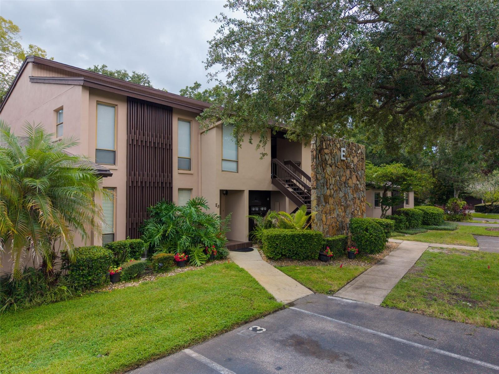Two-story beige condominium building with an exterior staircase, landscaping, and a stone entrance marker labeled E.