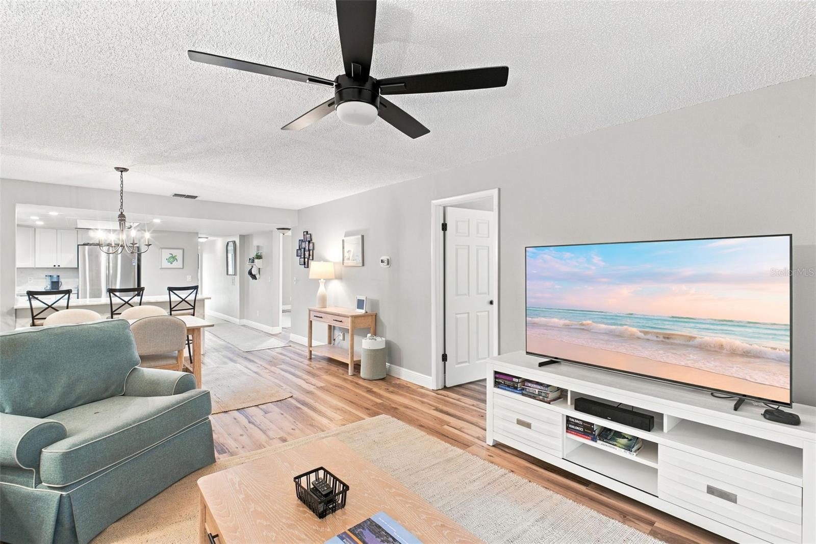 Living room with teal sofa, wood-look flooring, ceiling fan, and television beside an open dining area.