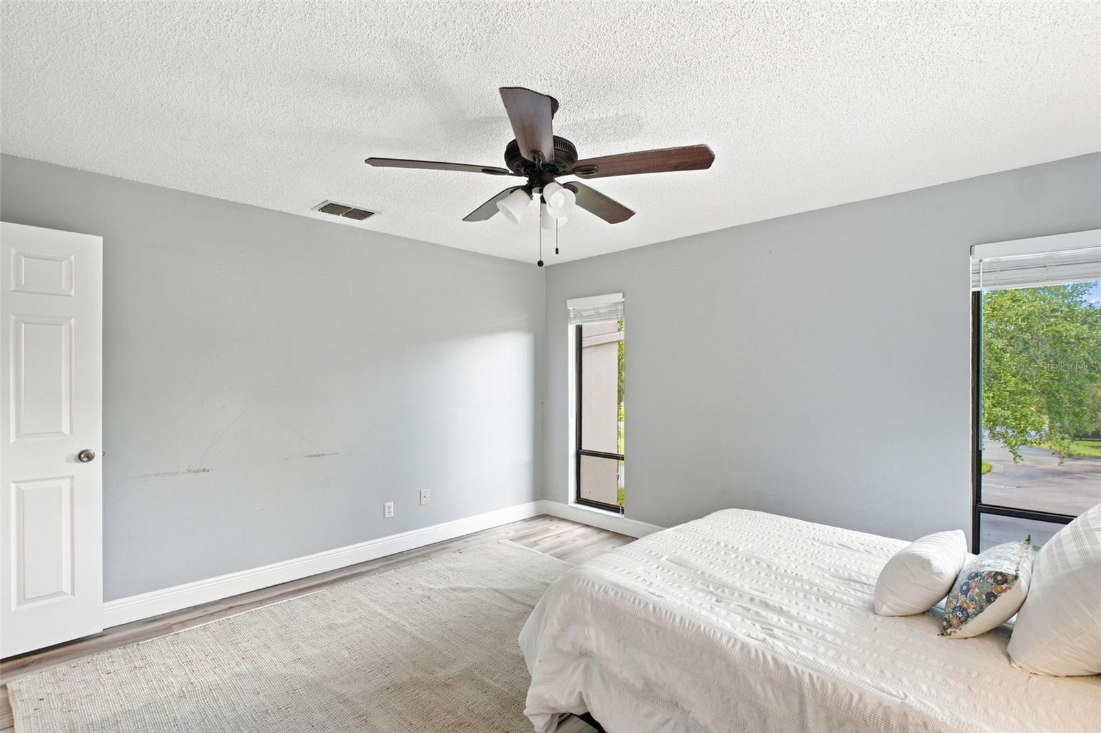 Bedroom with a white bed, gray walls, ceiling fan, and windows overlooking greenery.