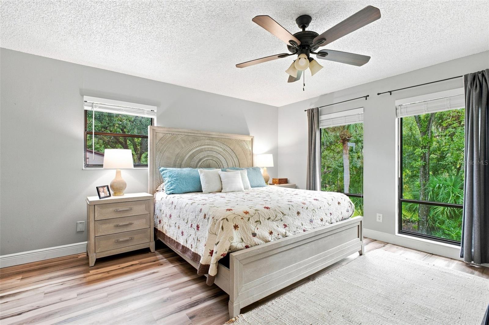 Staged bedroom with an arched carved headboard, floral quilt, and windows overlooking greenery.