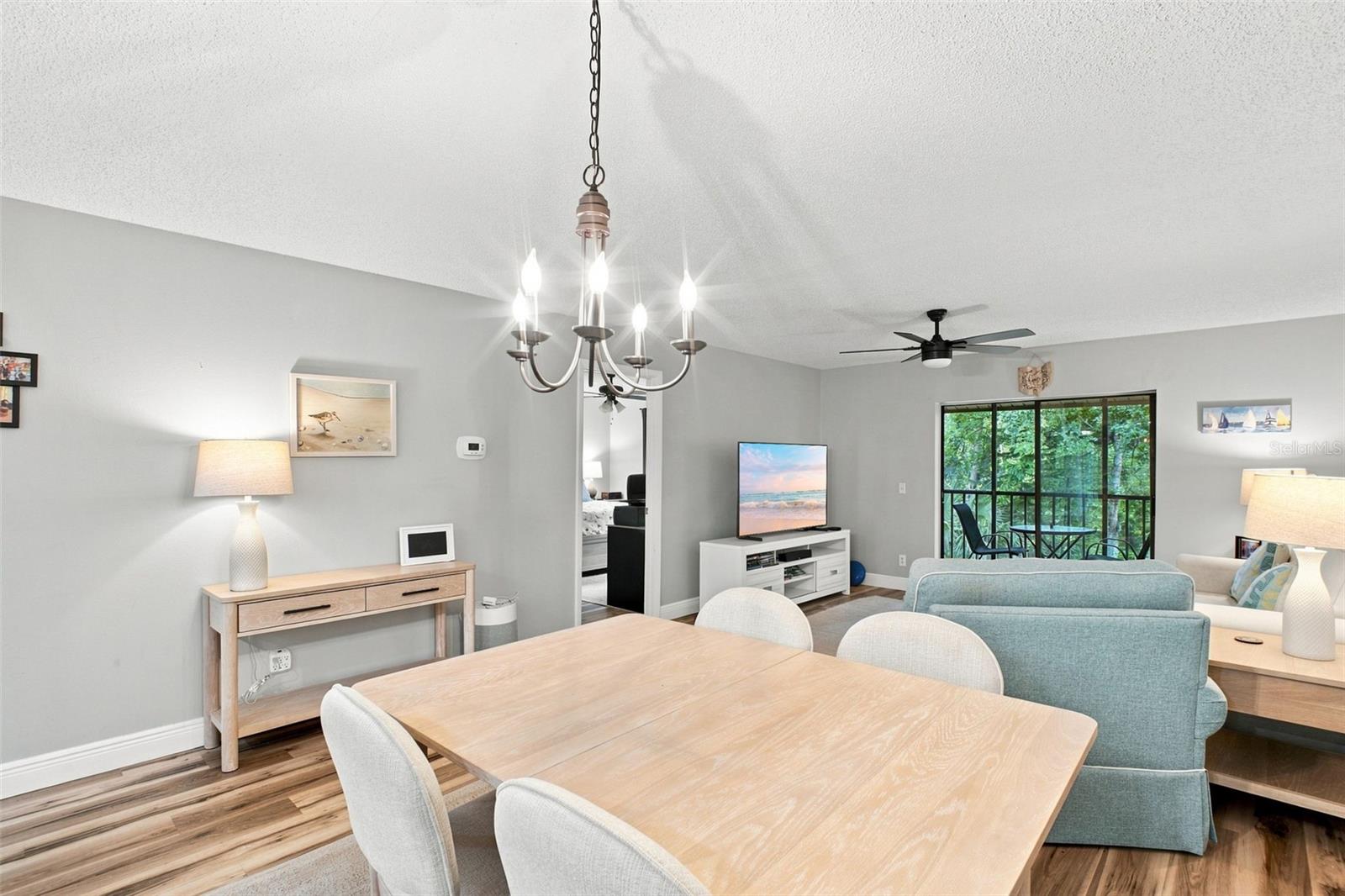 Furnished condo dining and living area with a light wood table, pale blue sofa, and leafy balcony view.