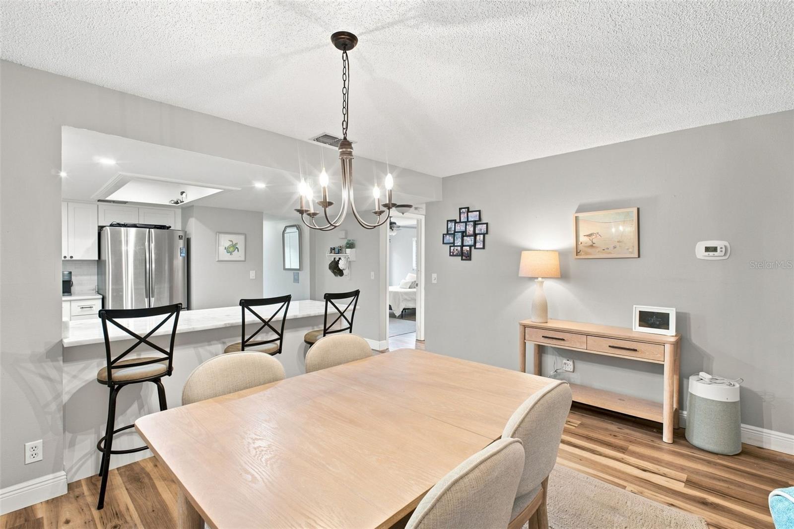 Dining area with a light wood table and chandelier beside an open kitchen with a breakfast bar.