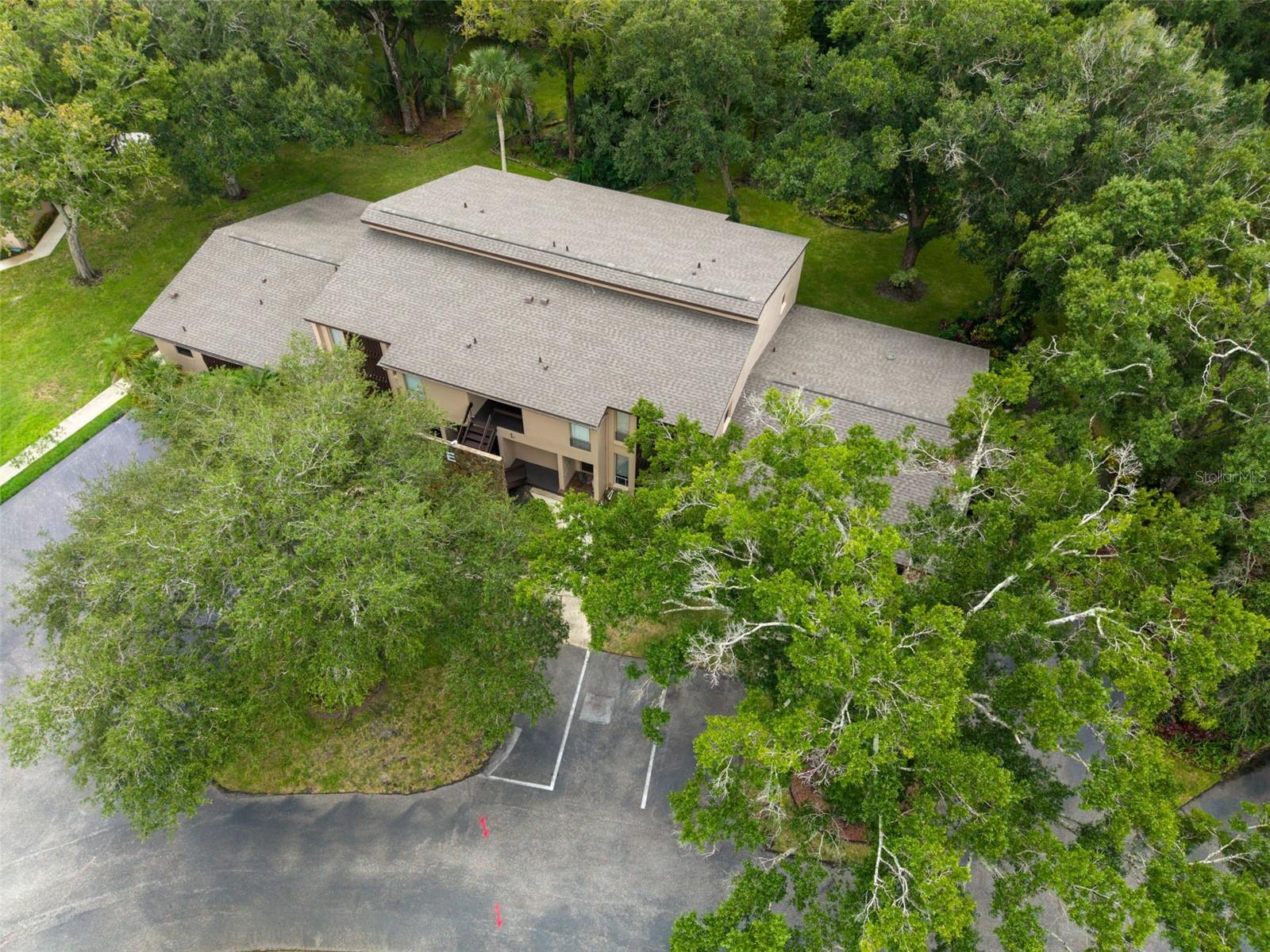 Aerial view of a beige residential building with broad shingle roofs, mature trees, and parking.