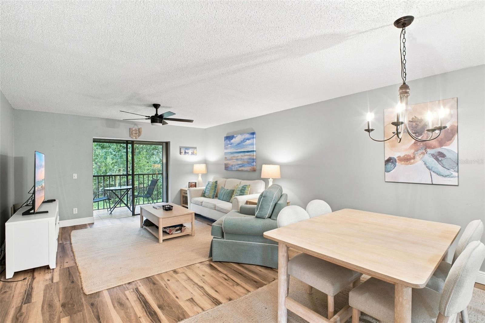 Furnished living and dining room with a teal armchair and balcony doors overlooking trees.
