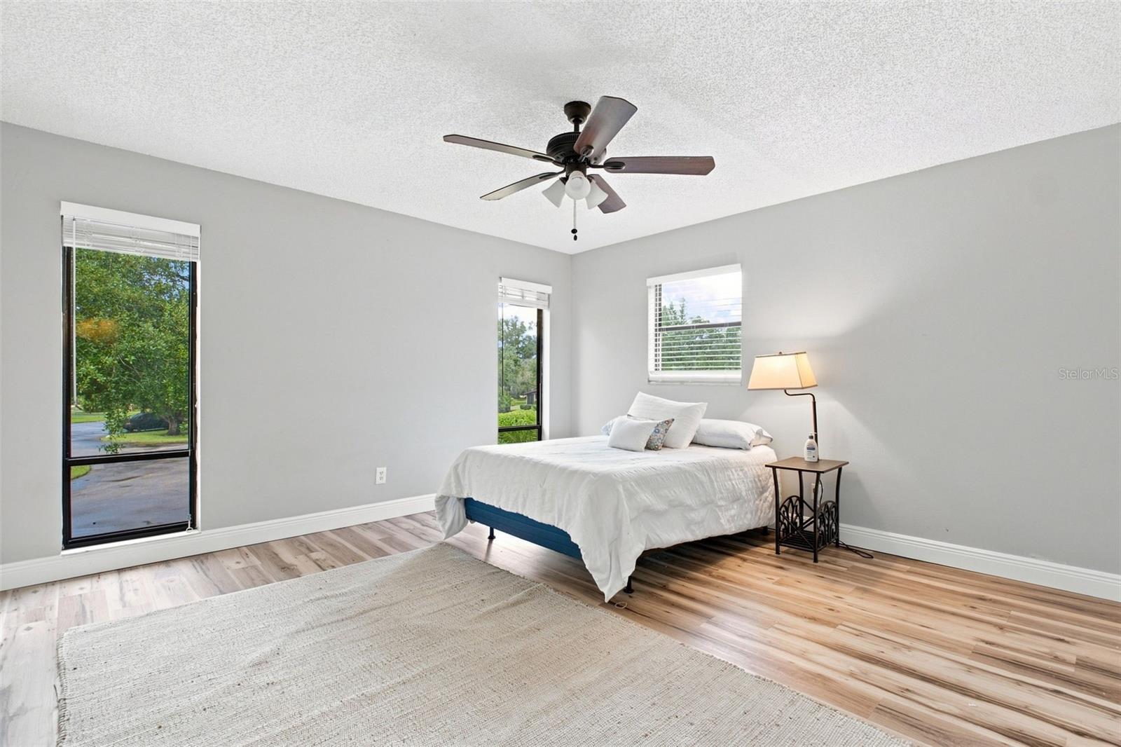 Bedroom with a white bed, ceiling fan, wood-look flooring, and tall windows.