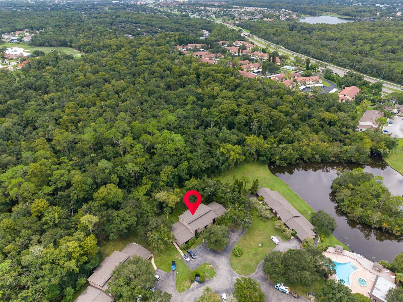 Aerial view of a marked condominium building among trees, with a pond and community pool nearby.