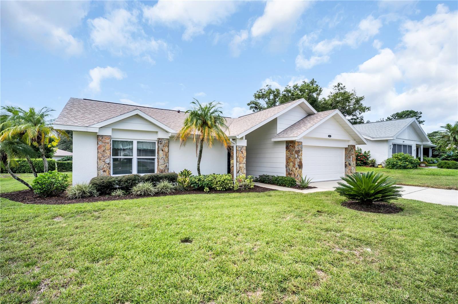 Single-story white home with stone accents, a two-car garage, palm trees, and a broad lawn.