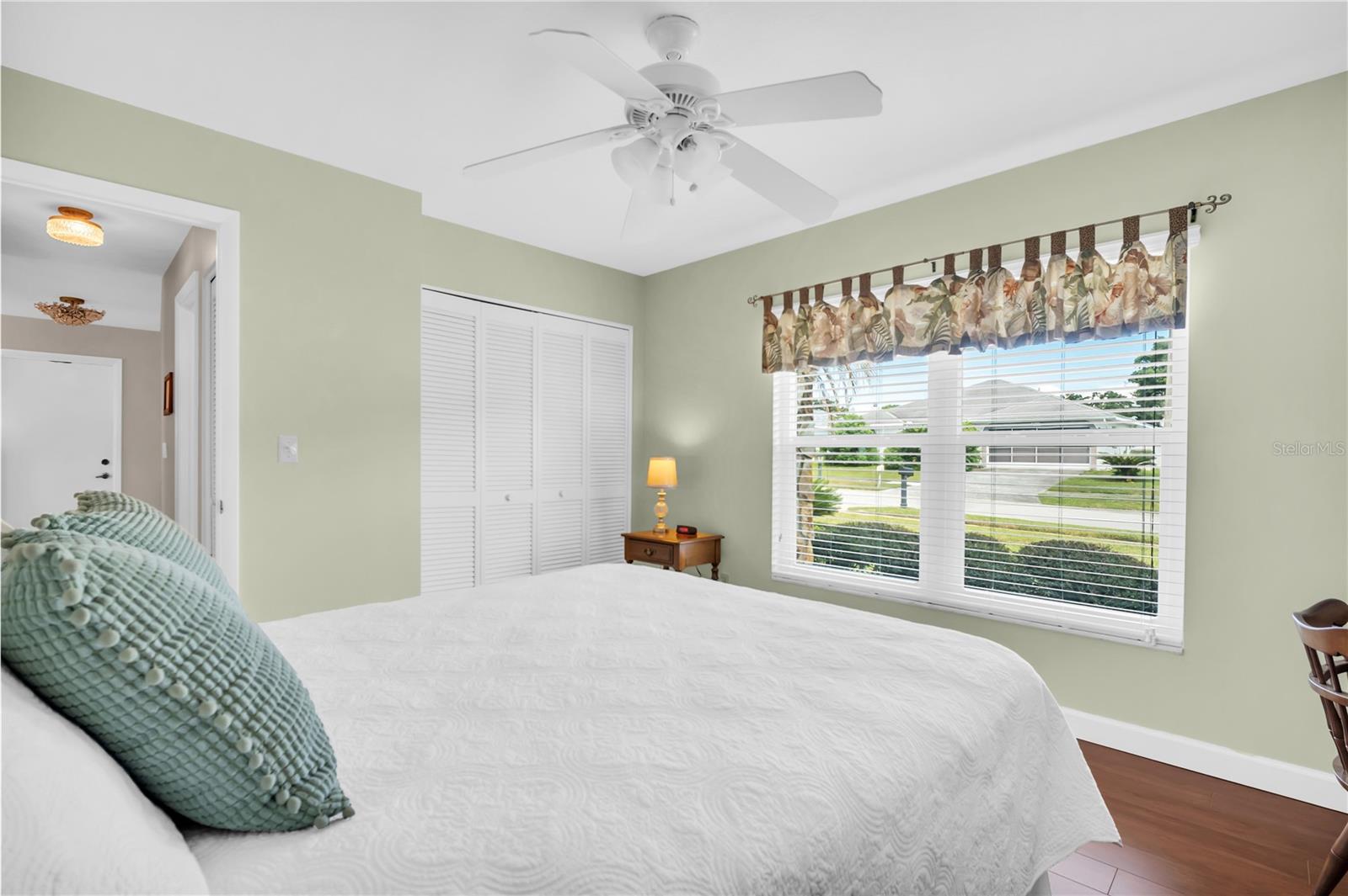 Bedroom with pale green walls, a white bed, louvered closet doors, and a large street-facing window.
