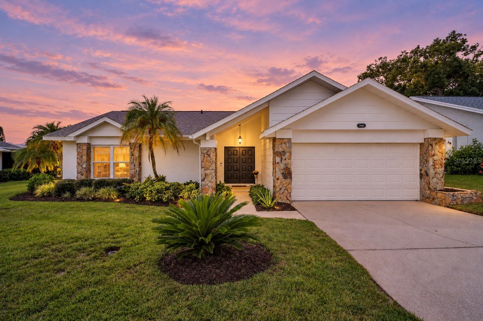 Single-story home with a two-car garage and tropical landscaping beneath a pink dusk sky.