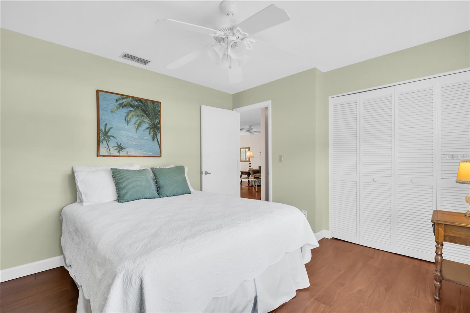Bedroom with a white bed, teal pillows, pale green walls, and framed tropical palm artwork.