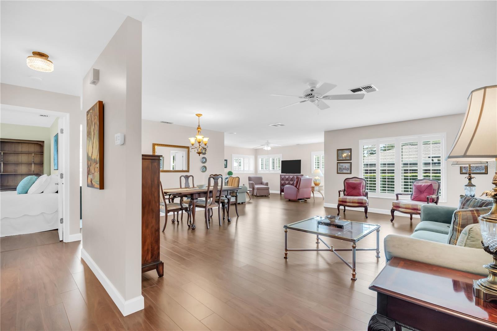 Open-plan living and dining room with plantation shutters, light wood floors, and a bedroom doorway.