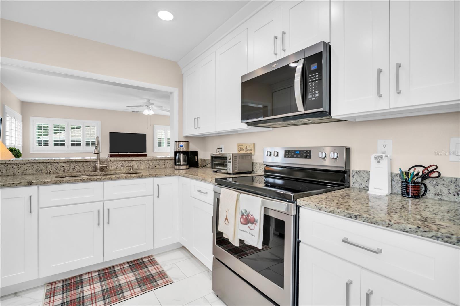 Bright kitchen with white cabinets, granite counters, and stainless steel appliances, opening to a living area.