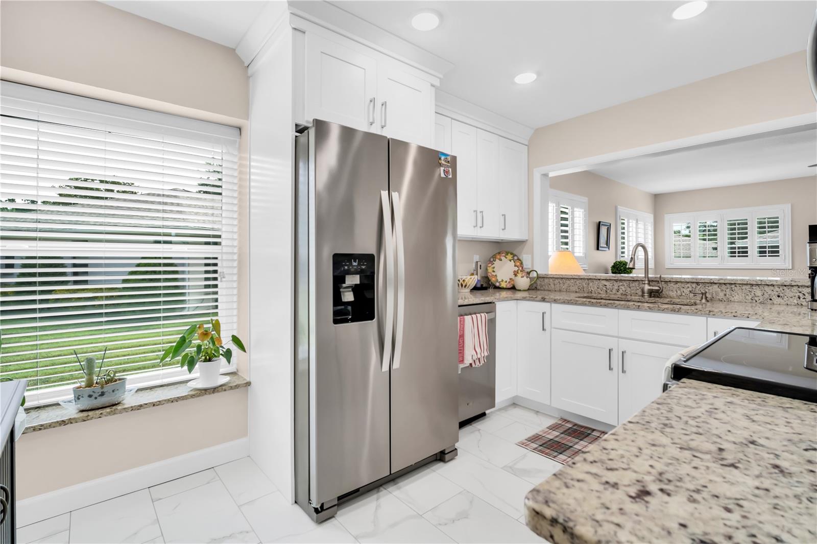 Kitchen with white cabinets, granite counters, stainless steel refrigerator, and a large window with blinds.