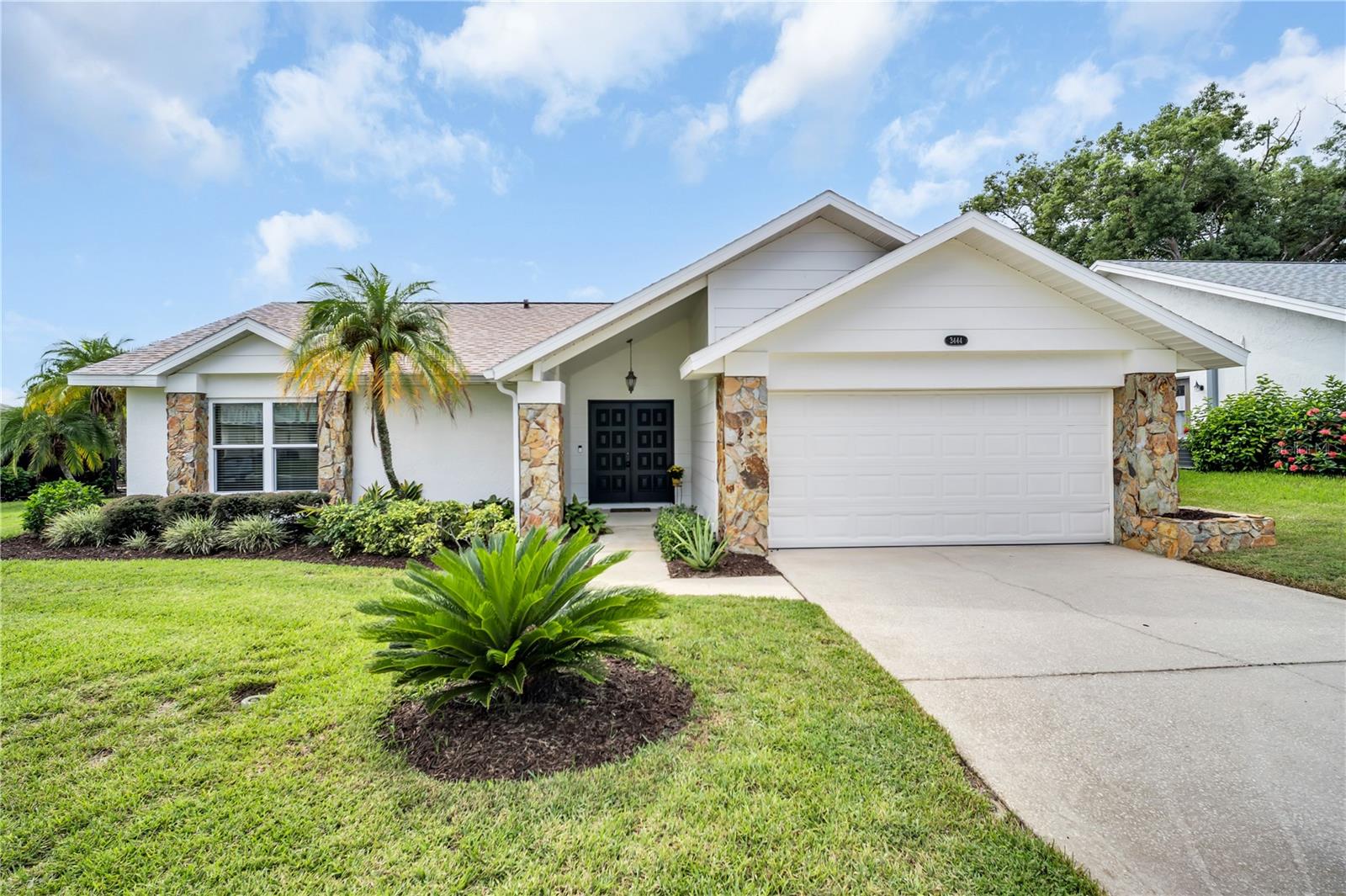 White single-story home with stone accents, a two-car garage, and palm trees in the landscaped yard.