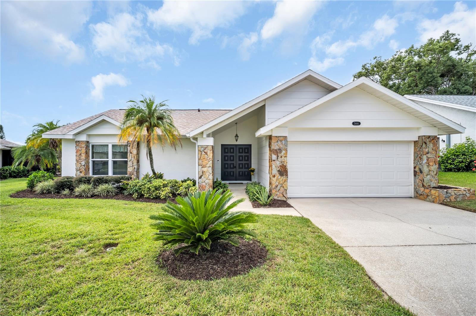 Single-story white home with a two-car garage, stone accents, and palm trees in the front yard.