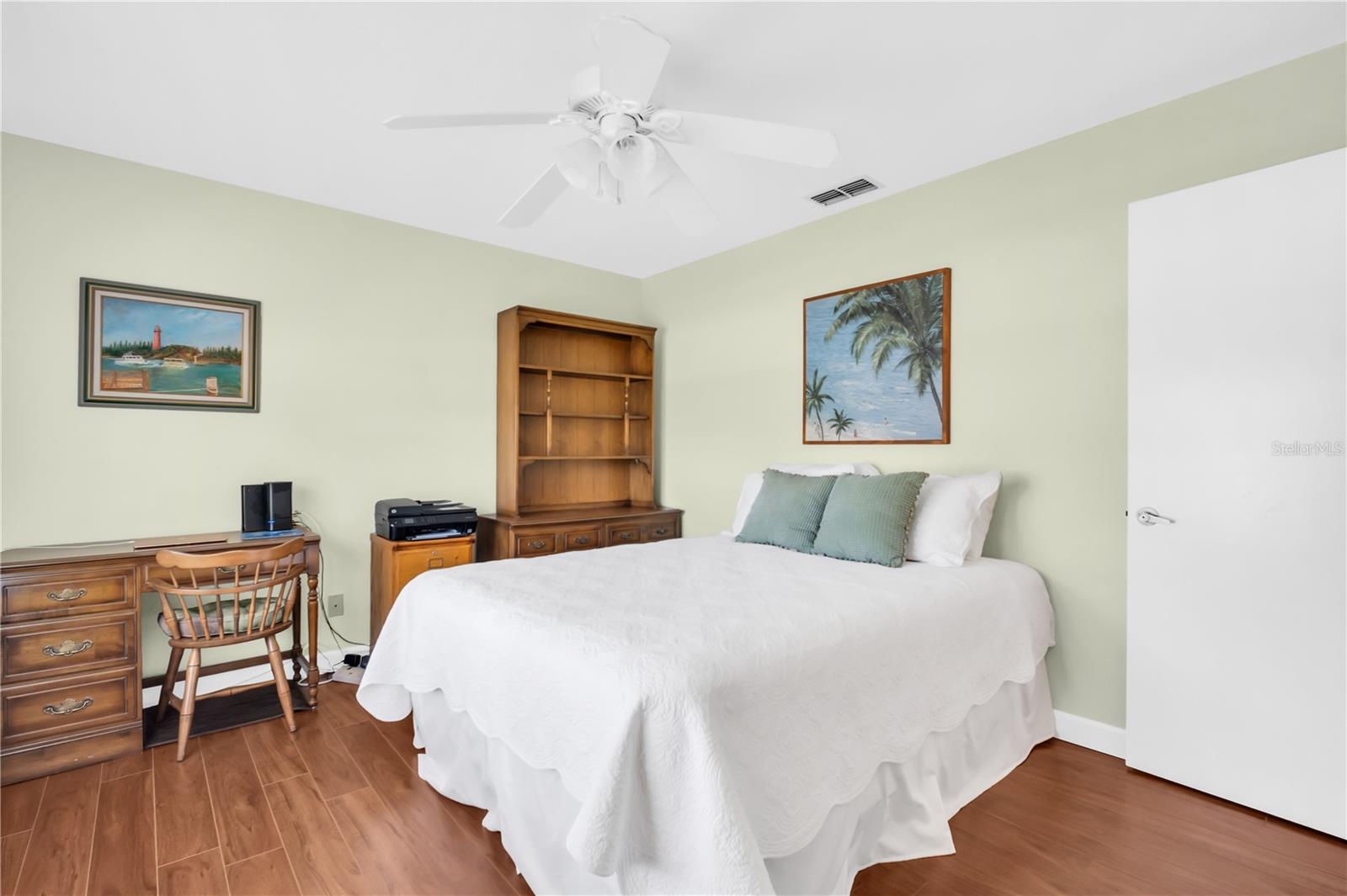 Furnished bedroom with a white bed, ceiling fan, and wooden desk and bookcase.
