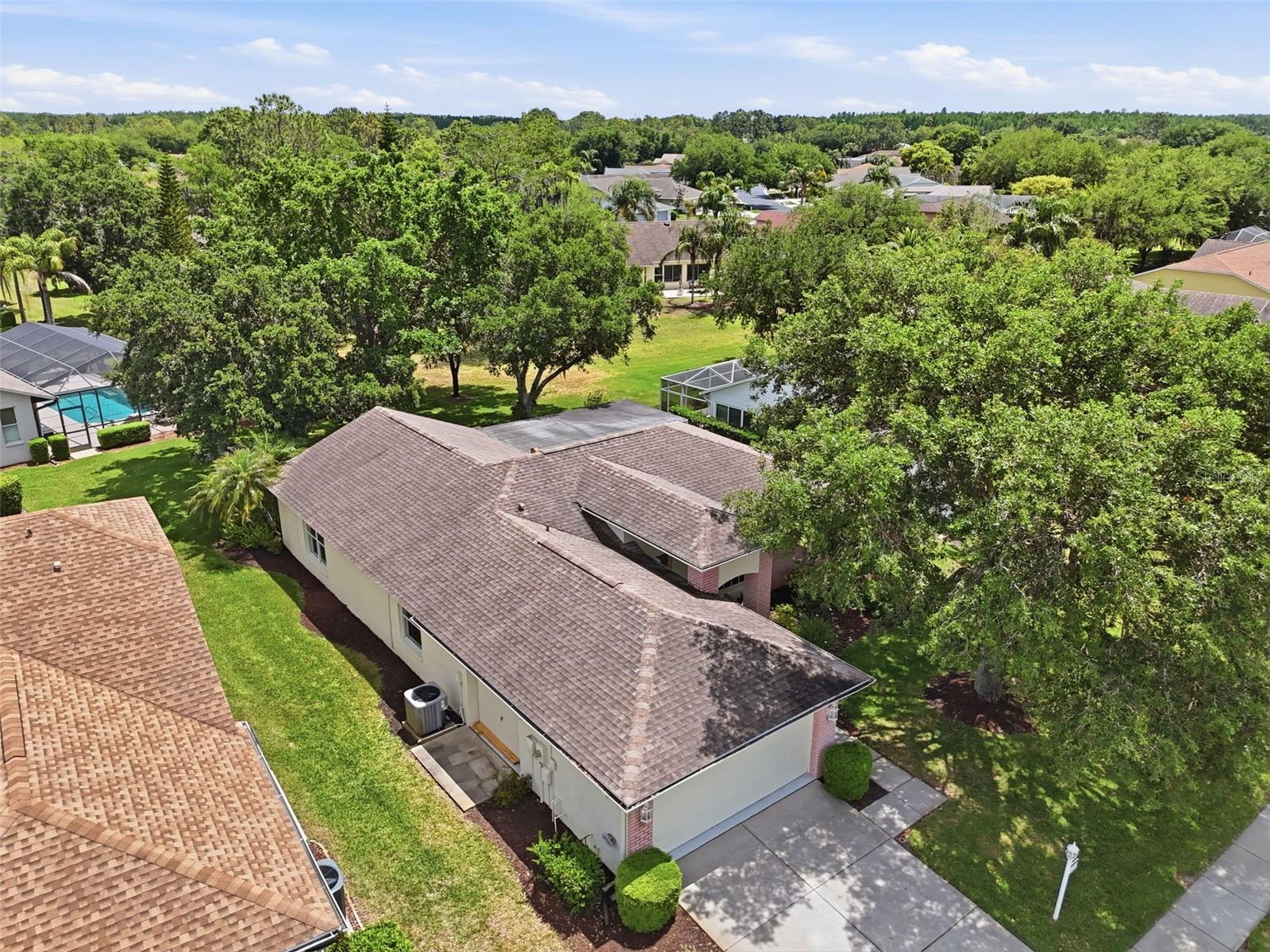 Aerial view of a single-story home with a broad shingle roof, driveway, and mature trees.