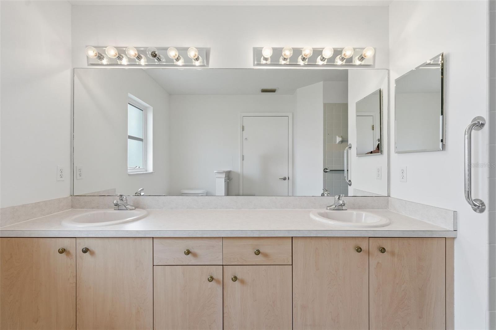 Bathroom vanity with two sinks, light wood cabinets, and a wide mirror.