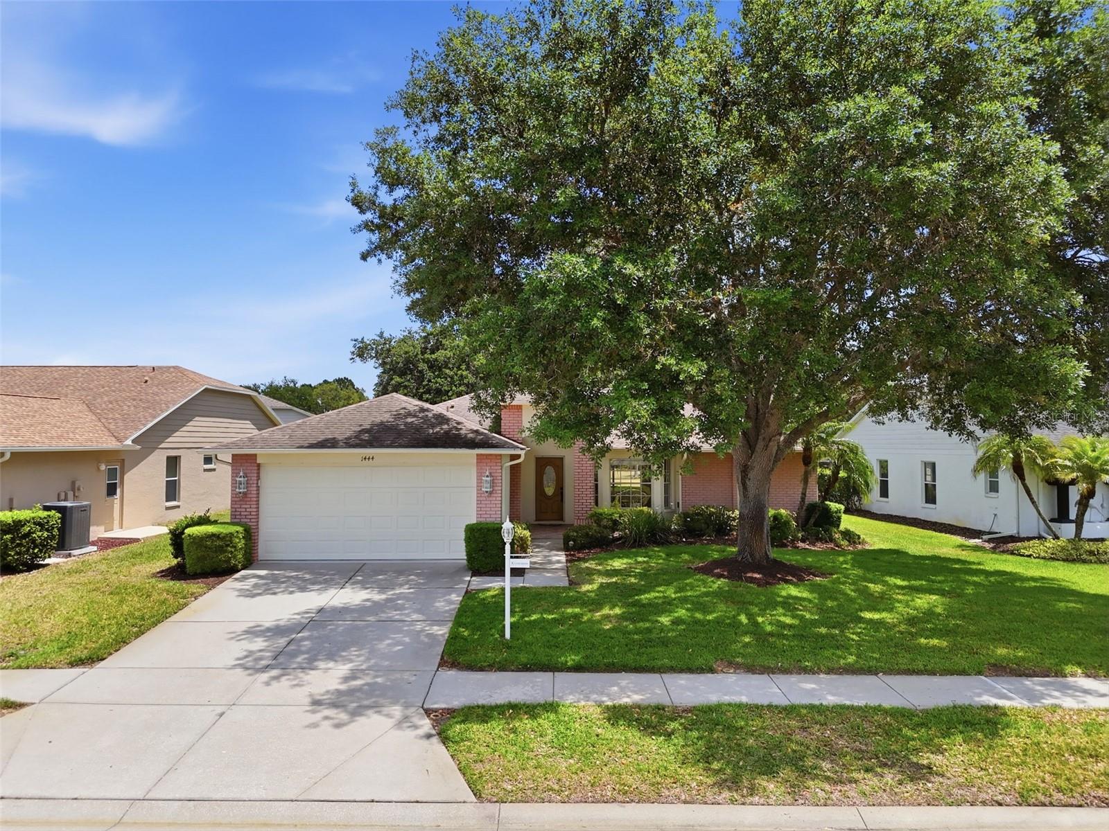 Single-story brick home with a two-car garage, broad driveway, and large shade tree on the front lawn.
