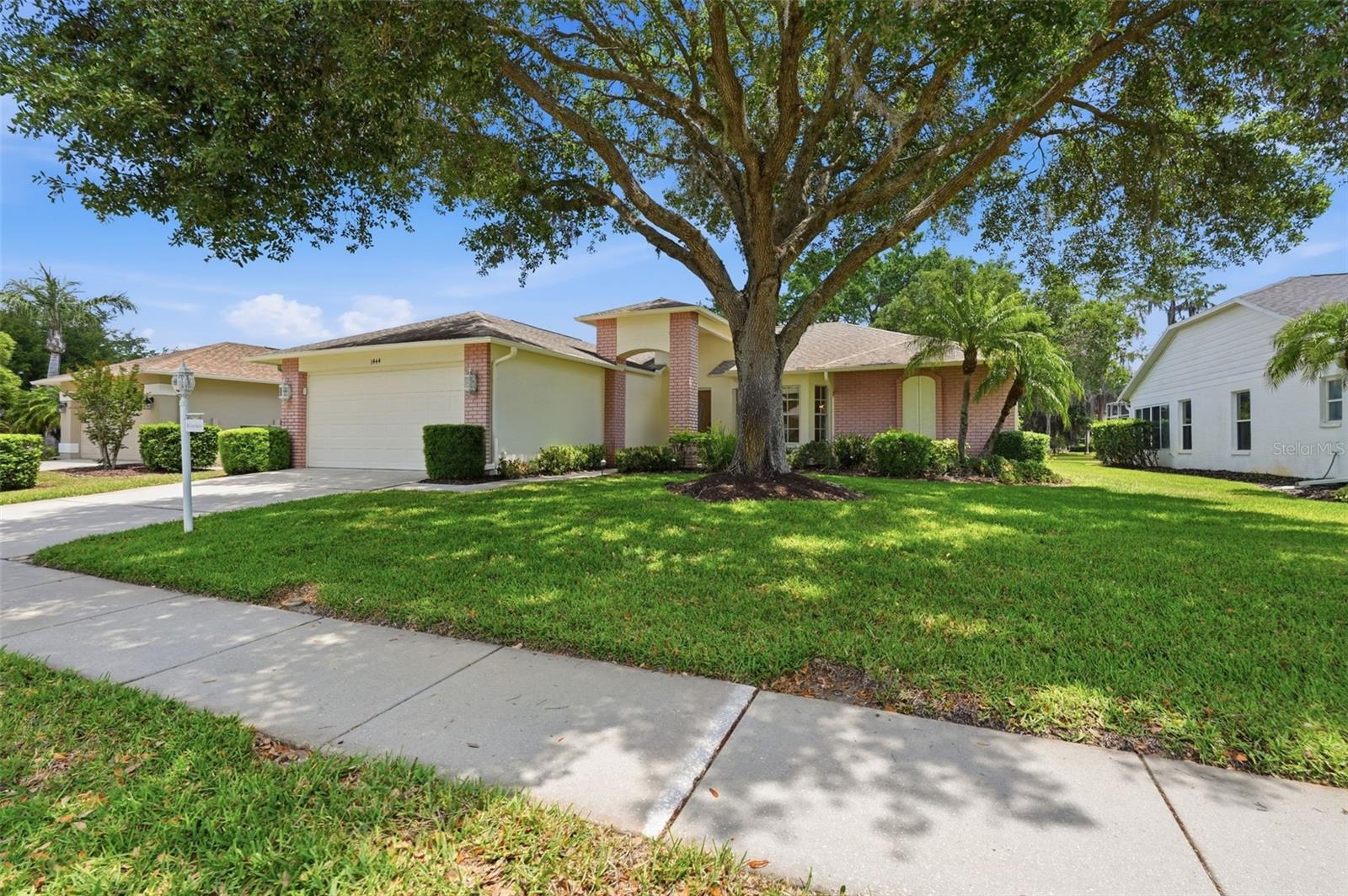 Single-story home with a two-car garage and brick accents beneath a large oak tree.