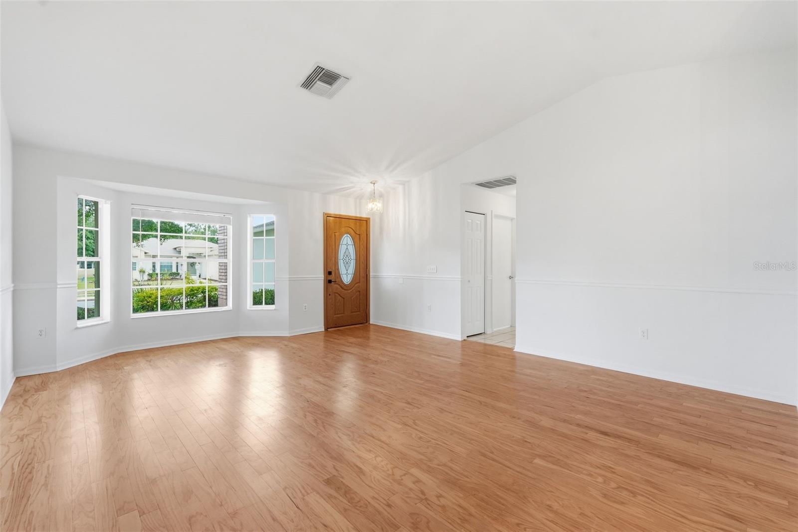 Empty living room with light wood floors, white walls, and a wooden front door beside large windows.