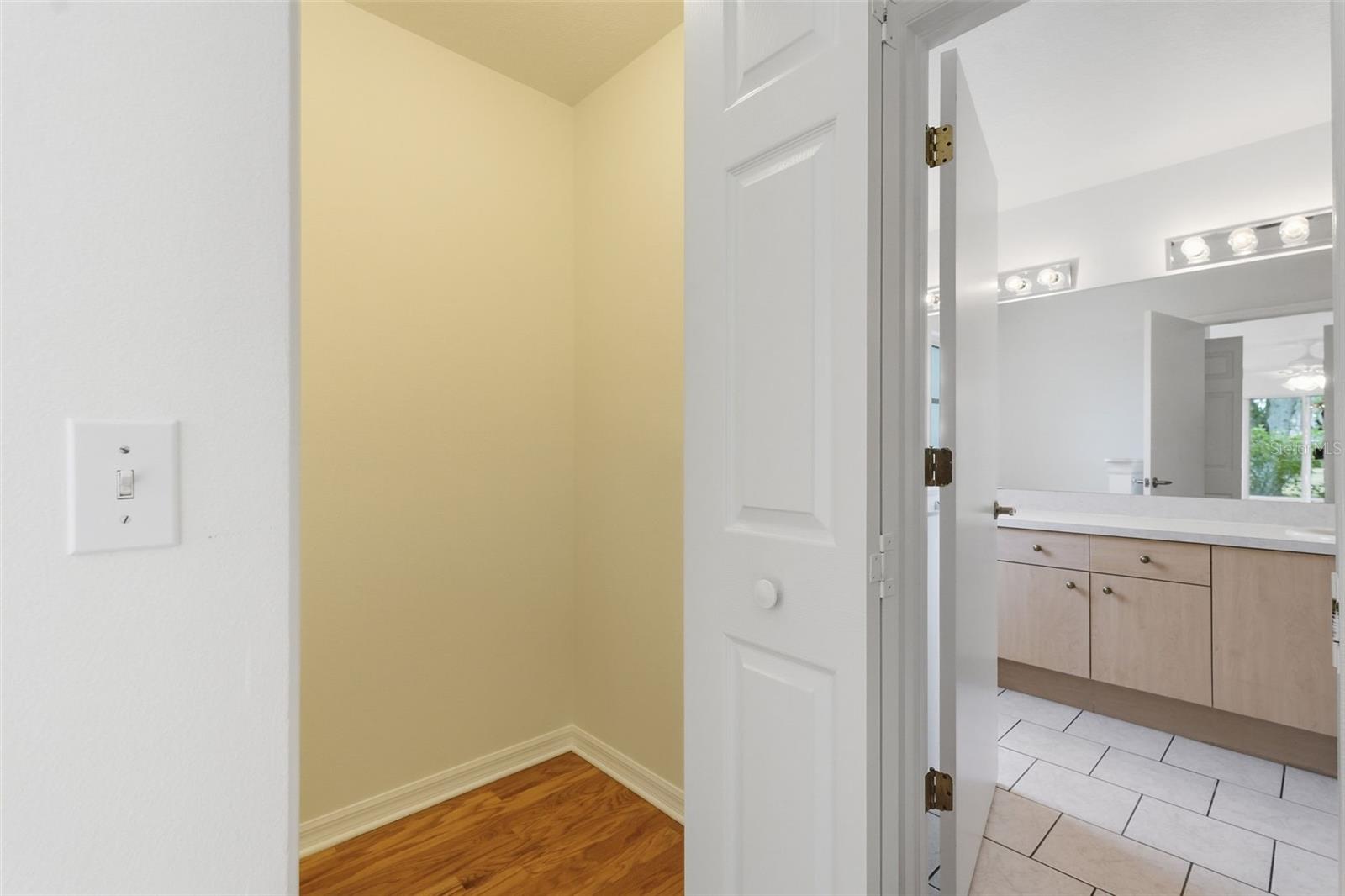 Bathroom with a light wood vanity and tiled floor, viewed past an open door beside a hardwood-floored nook.