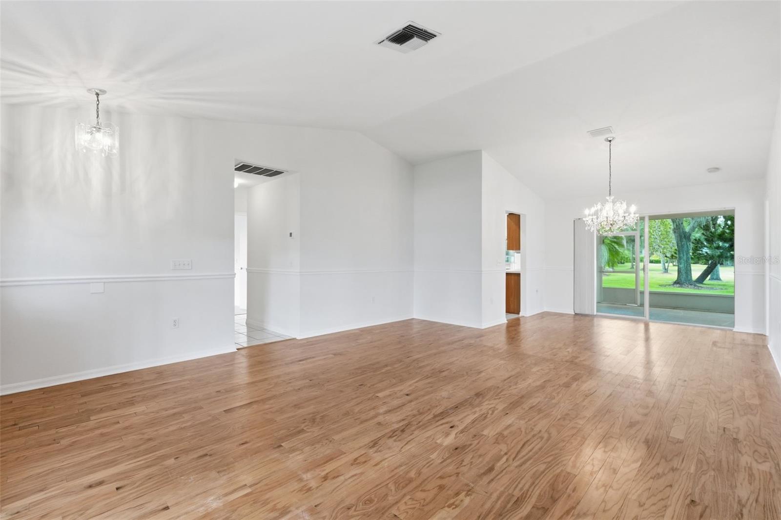 Vacant living and dining area with hardwood floors, two chandeliers, and sliding doors to a tree-lined lawn.