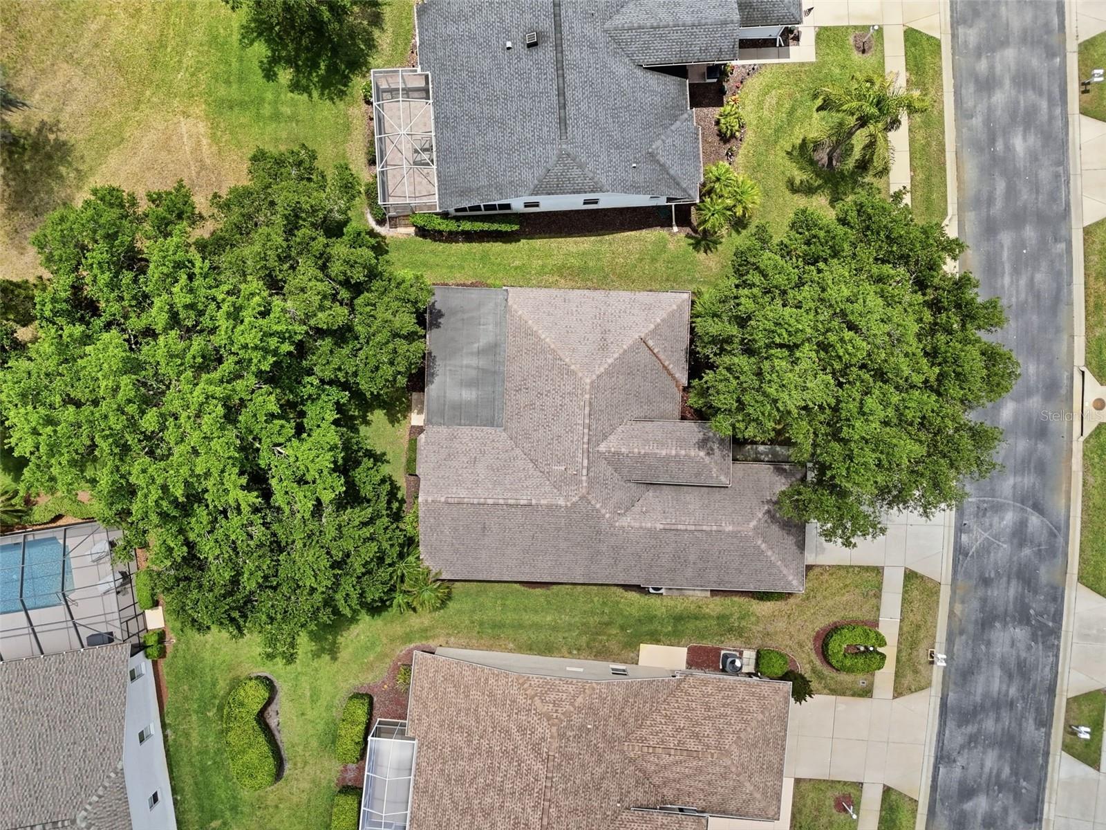 Aerial view of a home with a shingled roof, lawn, and mature trees beside a residential street.