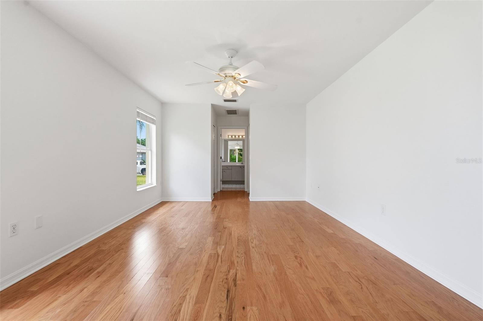 Empty bedroom with hardwood floors, ceiling fan, and doorway to a bathroom.