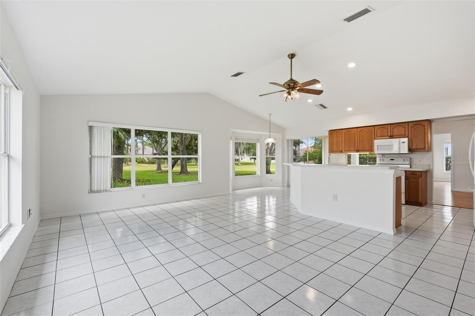 Open living and dining area with white tile floors, vaulted ceiling, and adjoining wood-cabinet kitchen.
