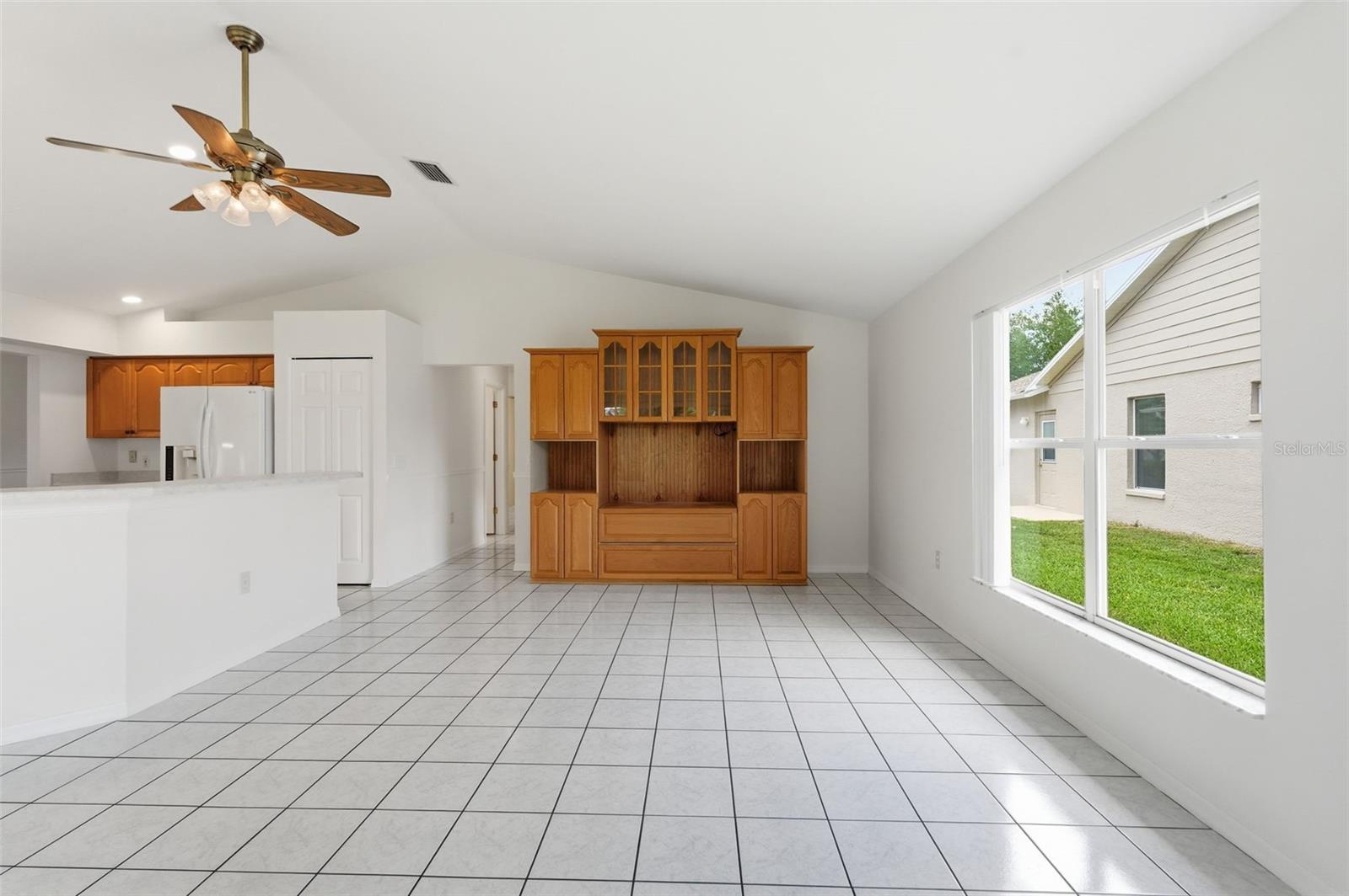 Open living area with tiled floors, vaulted ceiling, built-in wooden cabinet, and large window beside the kitchen.