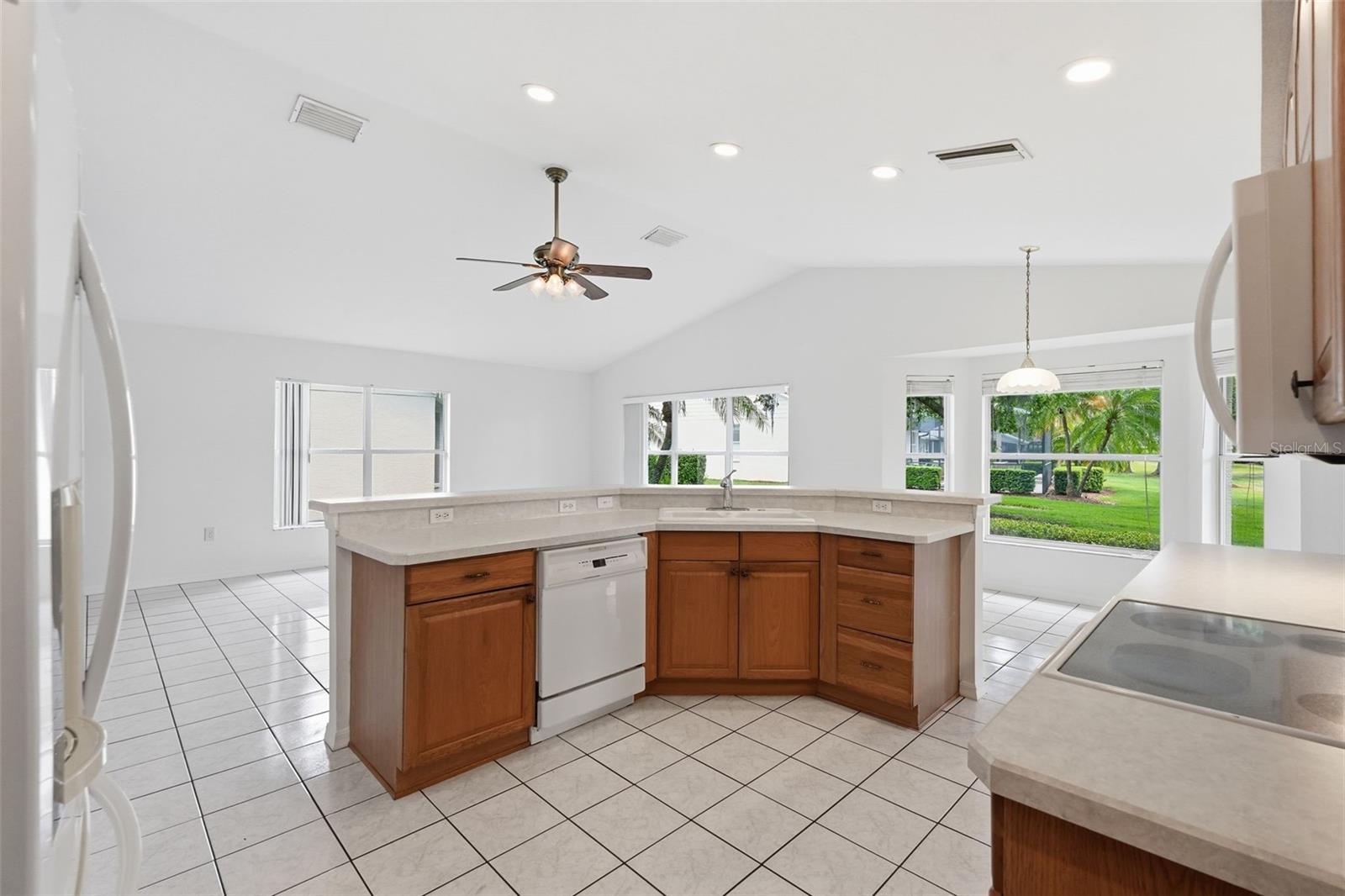 Bright kitchen with a wood-cabinet peninsula, white tile floors, vaulted ceiling, and windows overlooking a lawn.