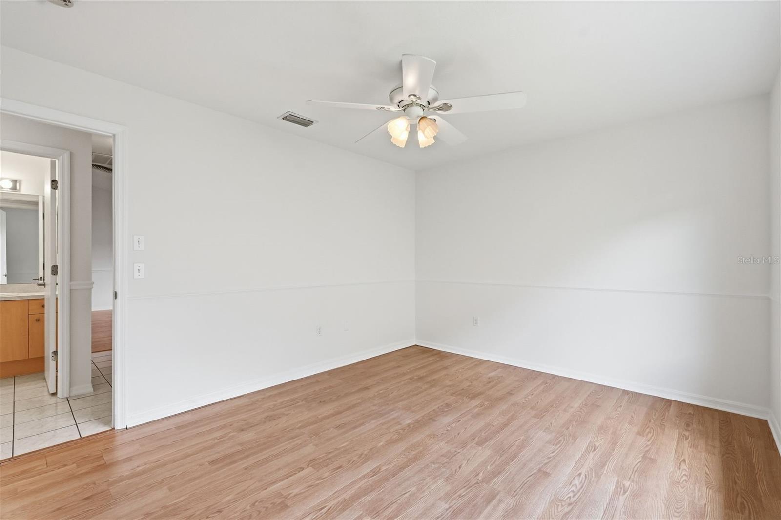 Empty bedroom with wood-look flooring, a ceiling fan, and a doorway to a tiled vanity area.