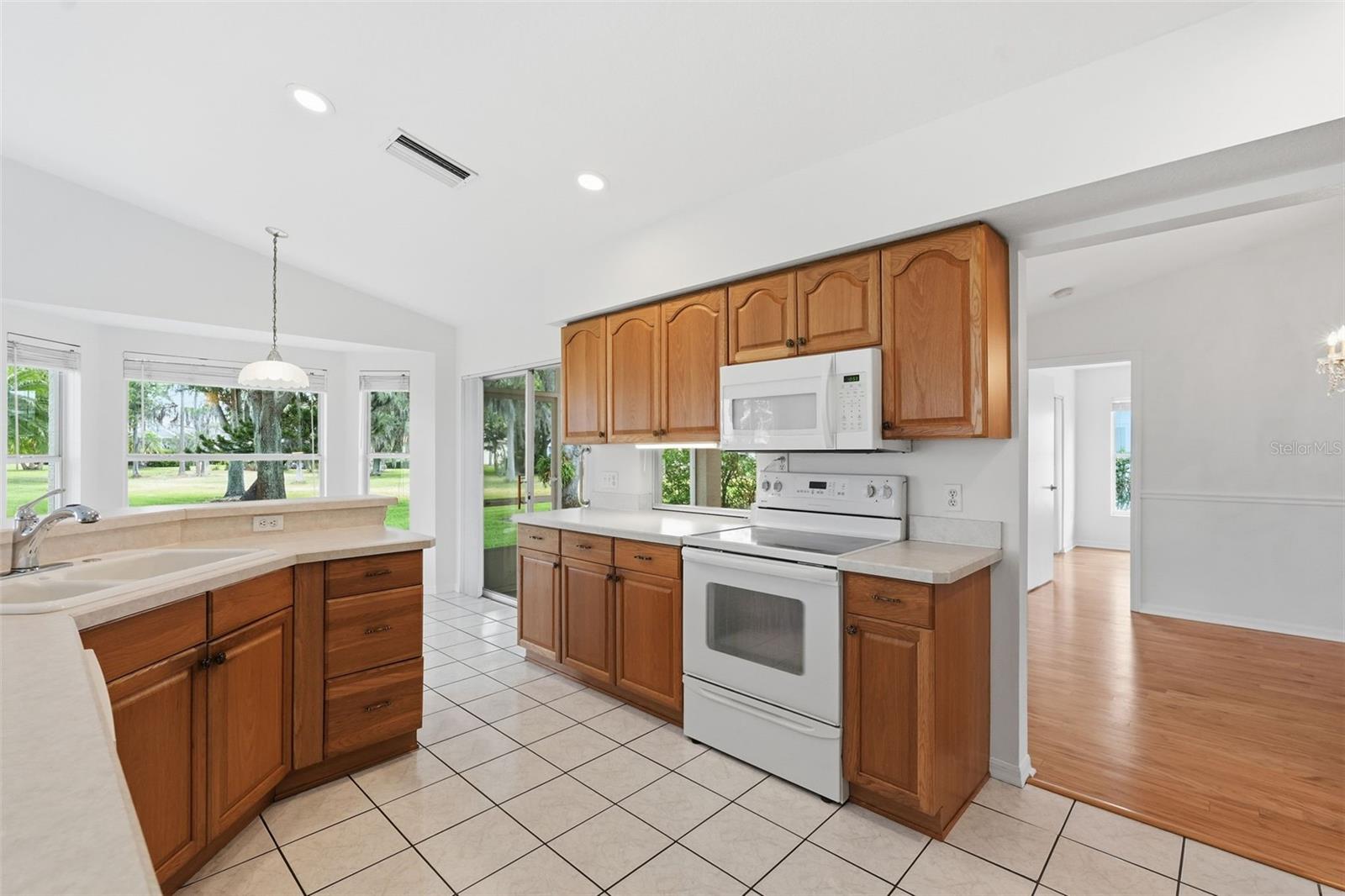 Kitchen with warm wood cabinets, white appliances, tiled floors, and windows overlooking a green yard.