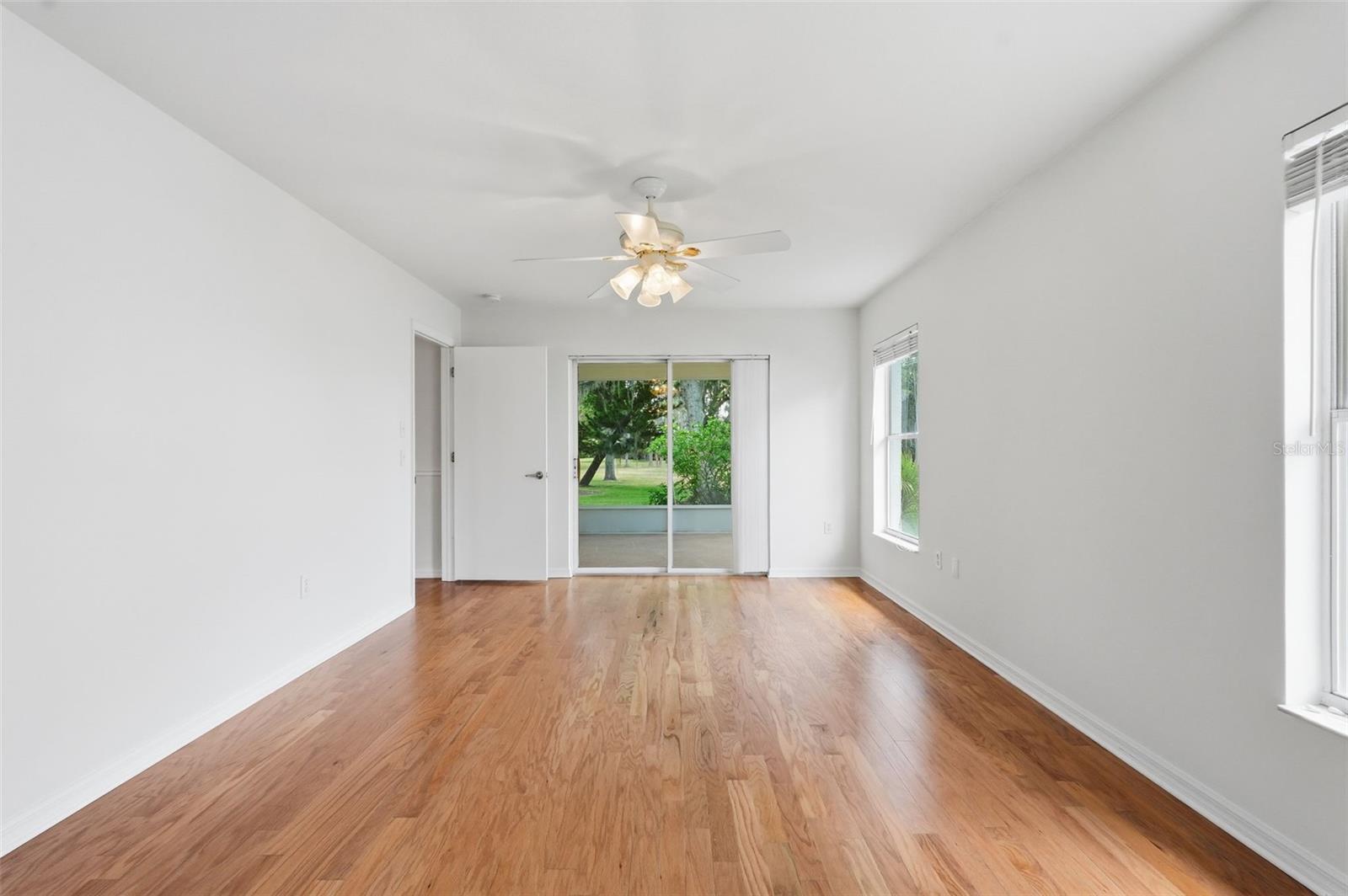 Empty room with hardwood floors, a ceiling fan, and sliding glass doors overlooking greenery.