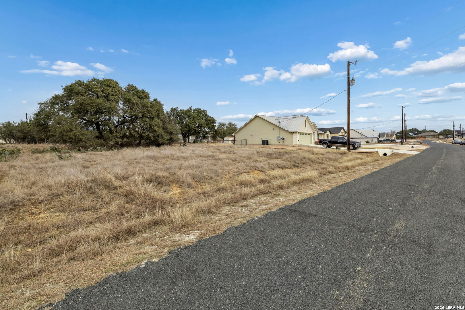 Vacant grassy lot beside a paved road, with mature oak trees and neighboring homes.