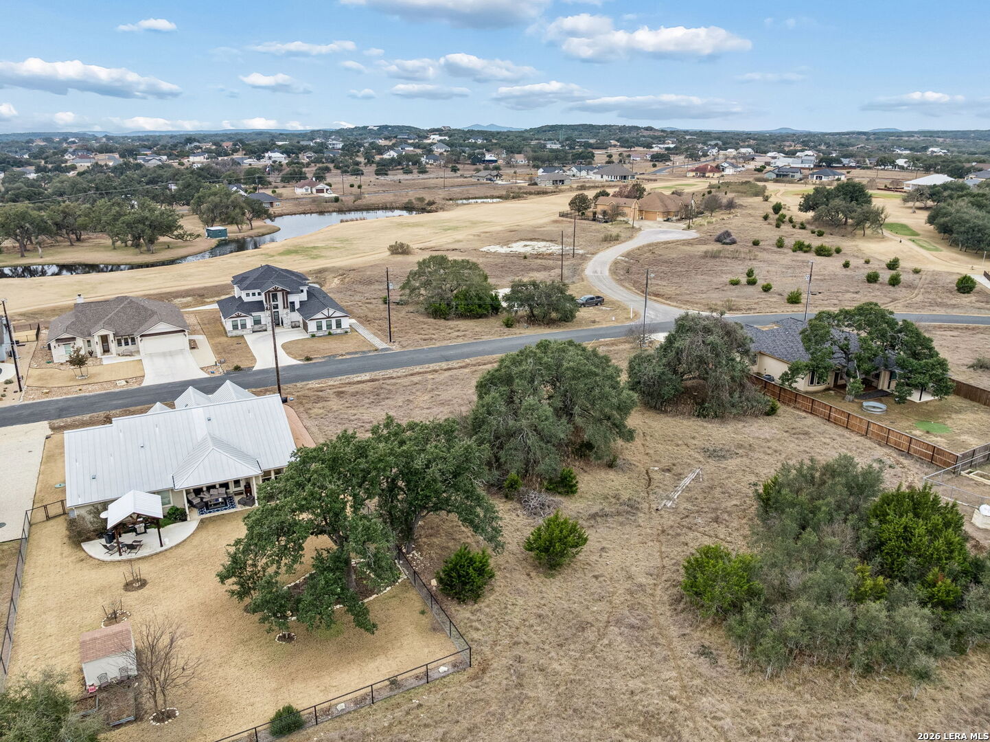 Aerial view of a dry residential lot with scattered trees, neighboring homes, and a small pond.