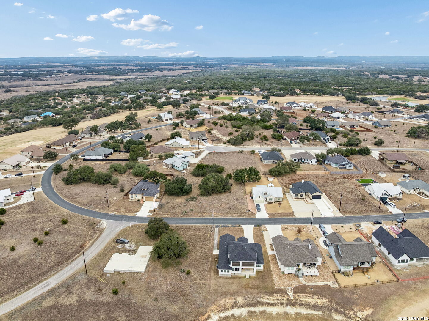 Aerial view of a residential neighborhood with curving roads, open lots, and low hills beyond.
