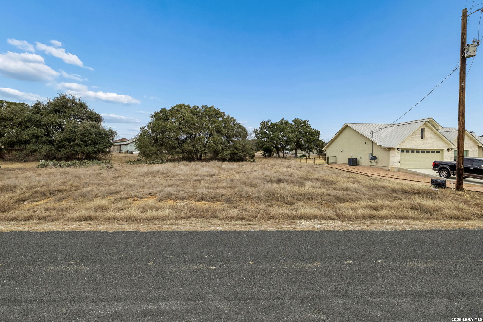 Vacant lot with dry grass and scattered trees beside a neighboring house.