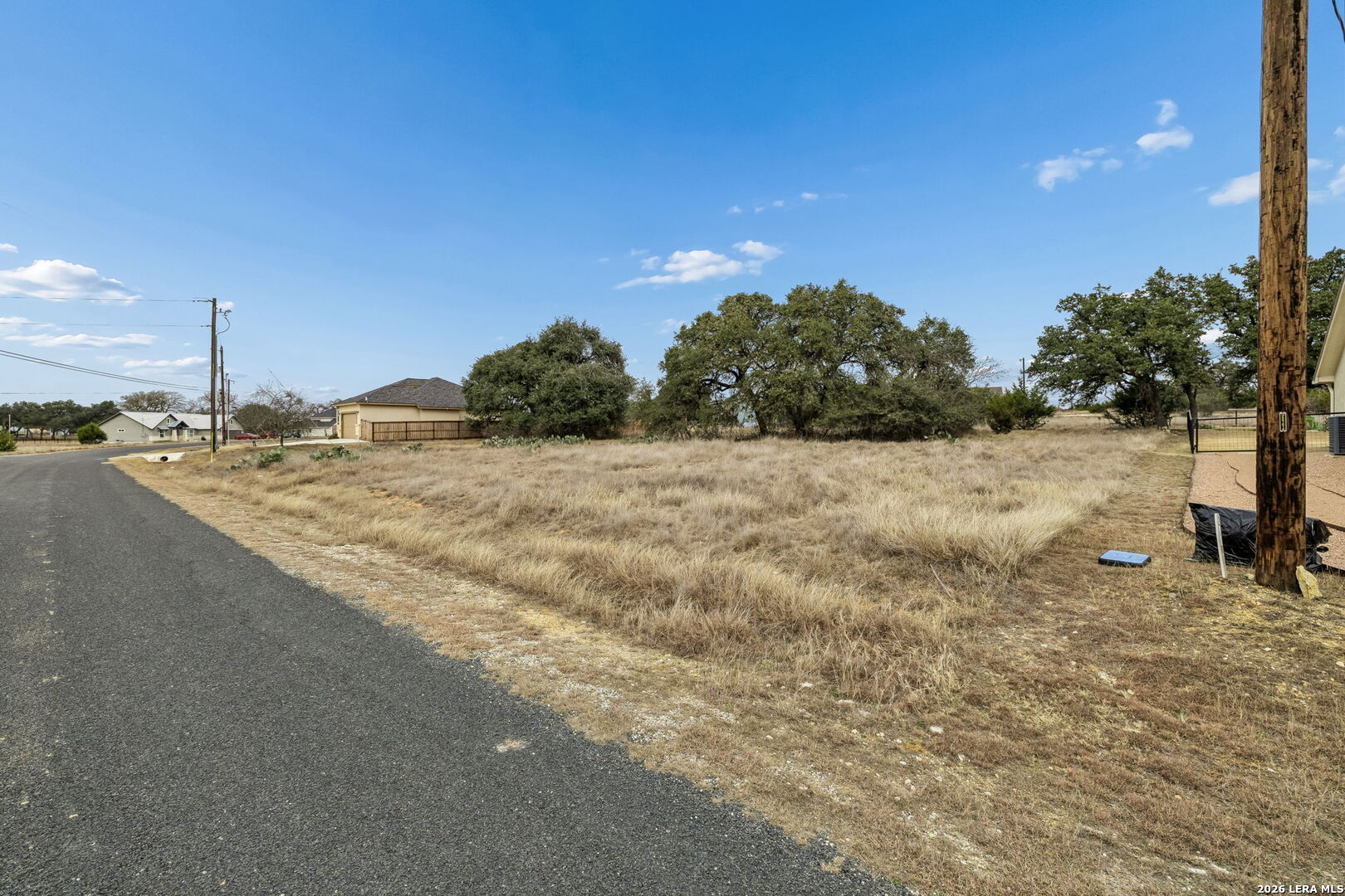 Vacant grassy lot beside a paved road, with mature oak trees and neighboring houses.
