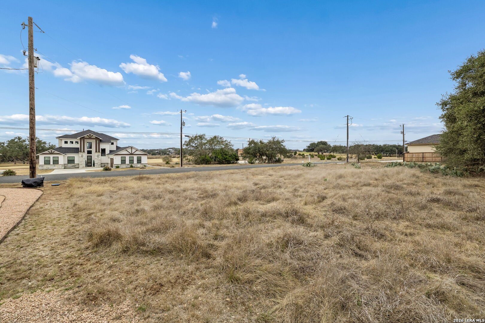 Vacant grassy lot across from a modern two-story home, with a road and utility poles.