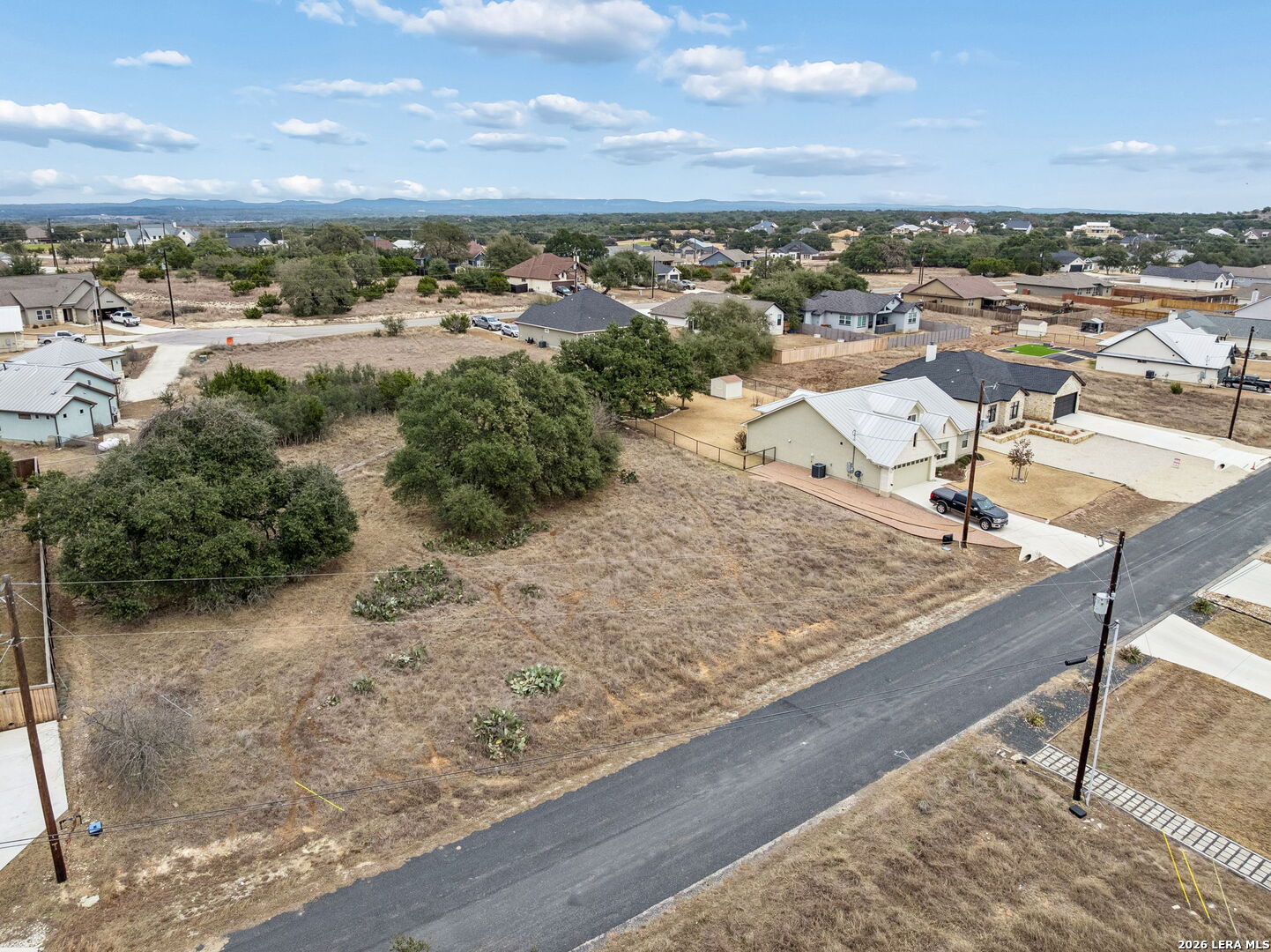 Vacant lot beside a paved road, surrounded by homes and trees.