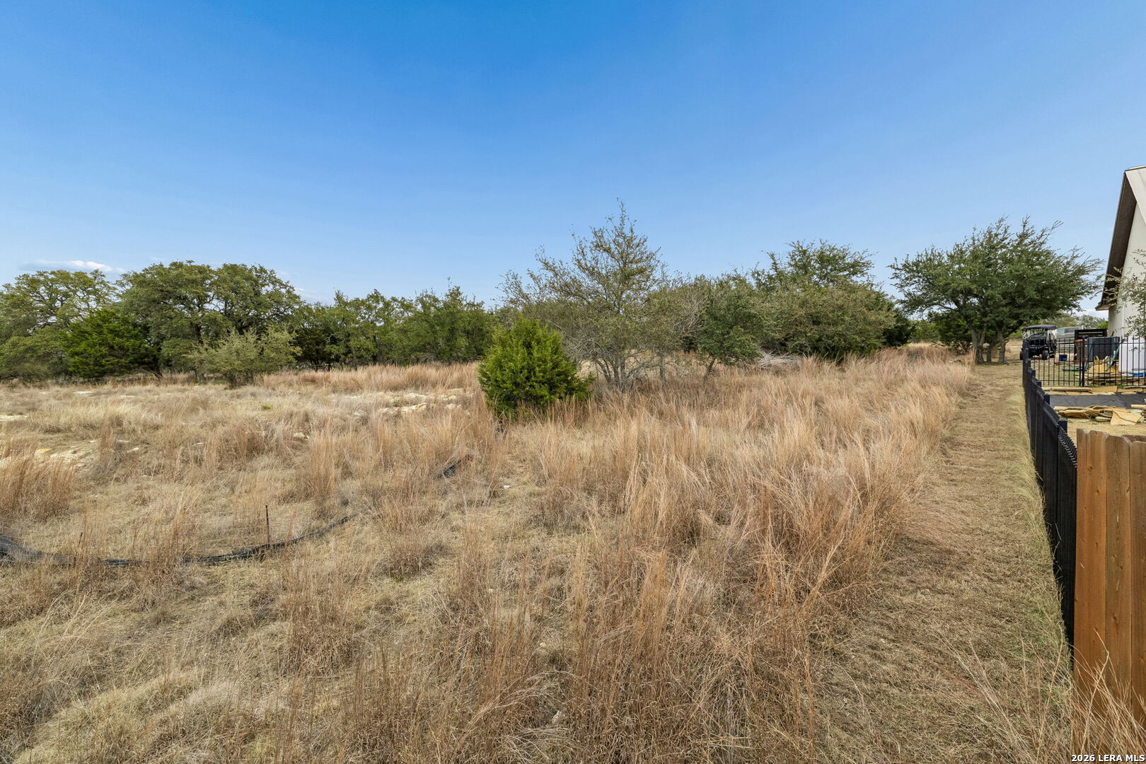 Grassy lot with scattered trees and brush beside a fenced path.