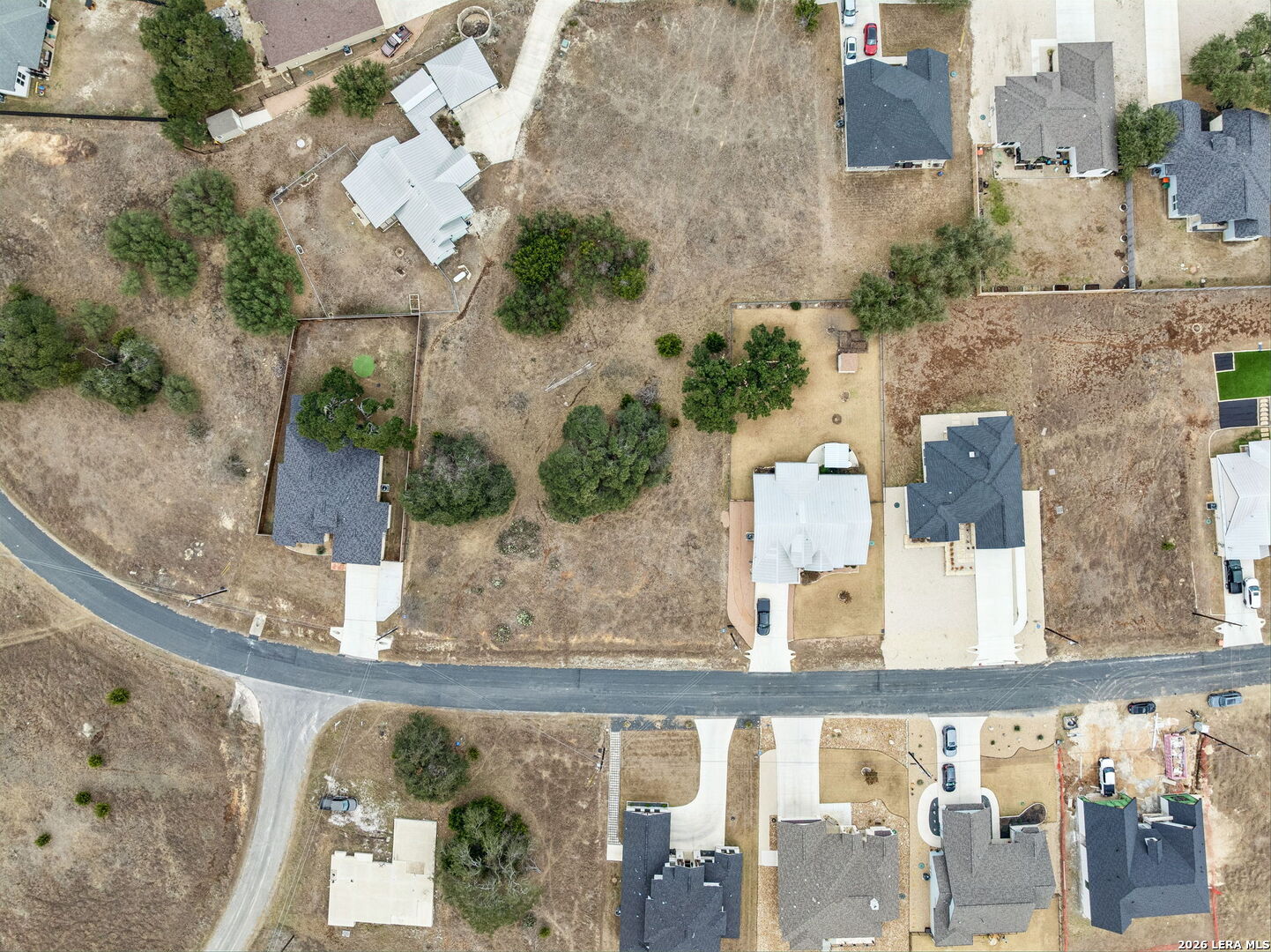 Aerial view of an open lot with scattered trees, neighboring homes, and a paved road.