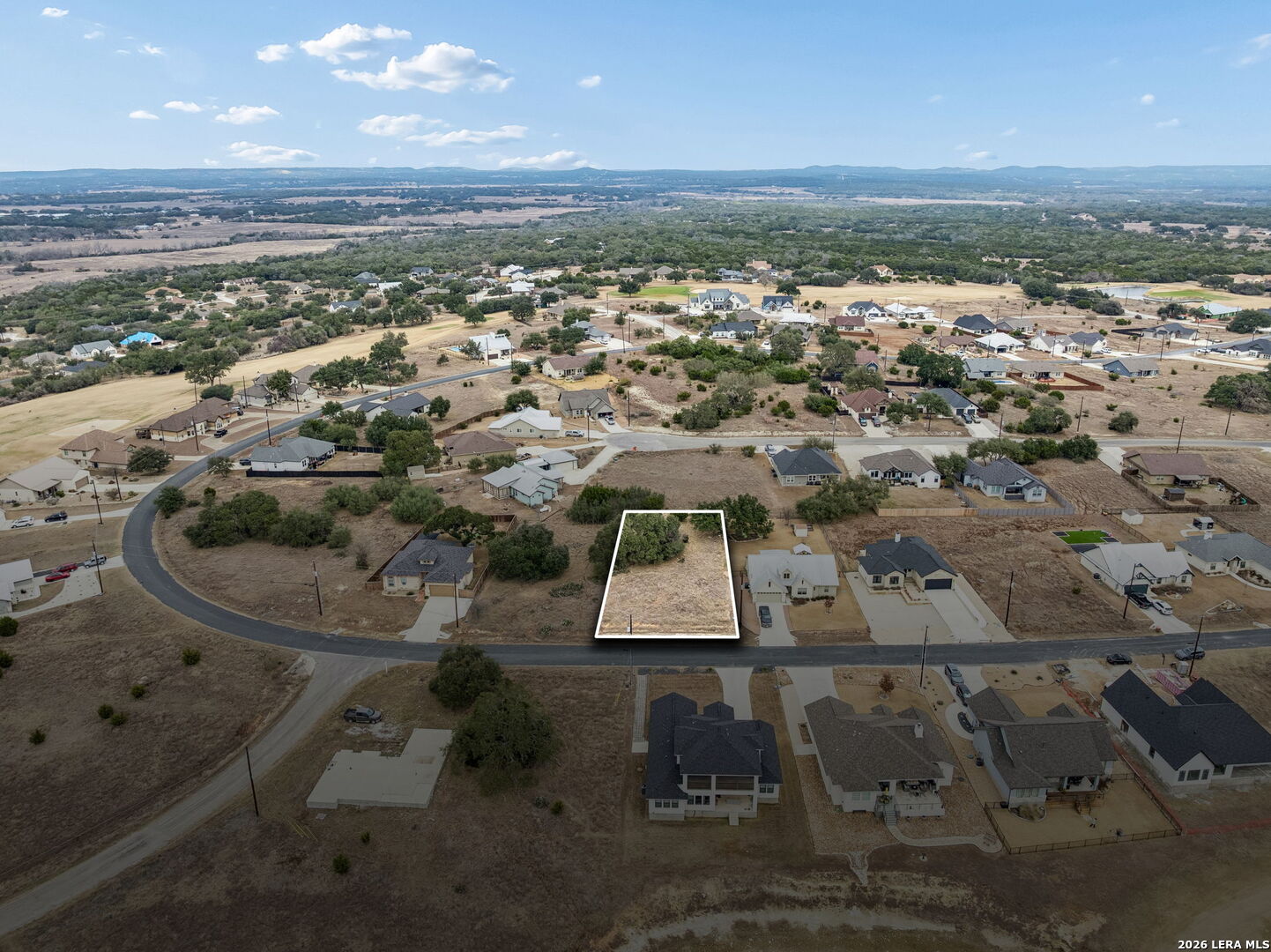 Vacant lot outlined in white among homes, with wooded countryside and distant hills beyond.