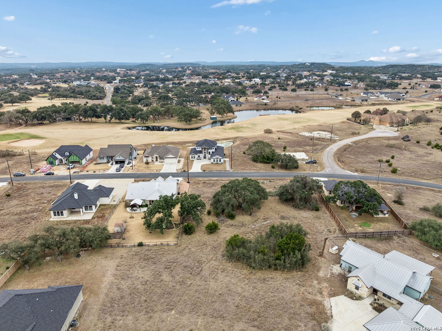 Aerial view of a dry neighborhood with scattered homes, an open lot, and a small pond.