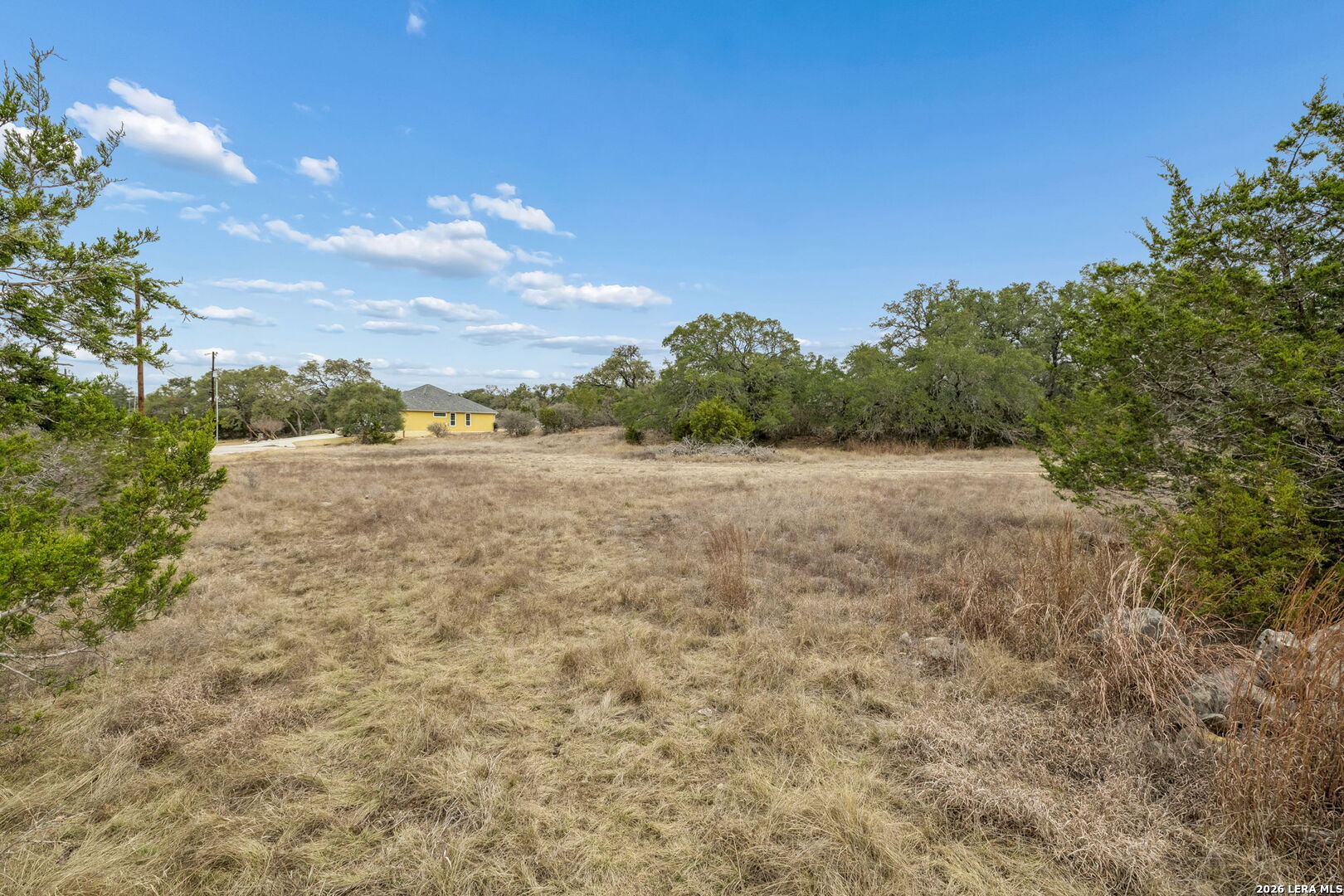 Open grassy lot bordered by trees, with a small yellow house in the distance.