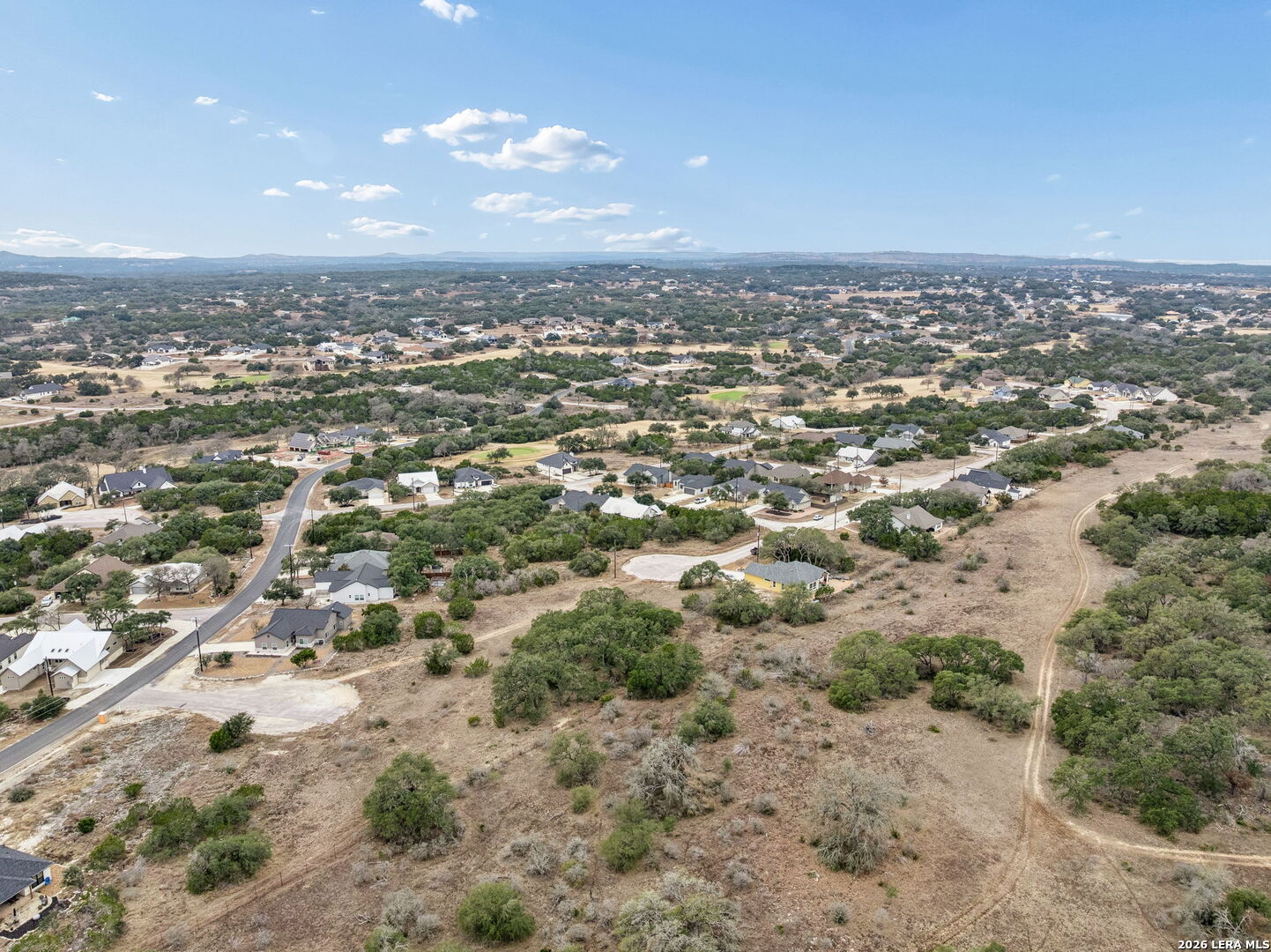 Aerial view of homes and roads beside brush-covered open land, with distant low hills.