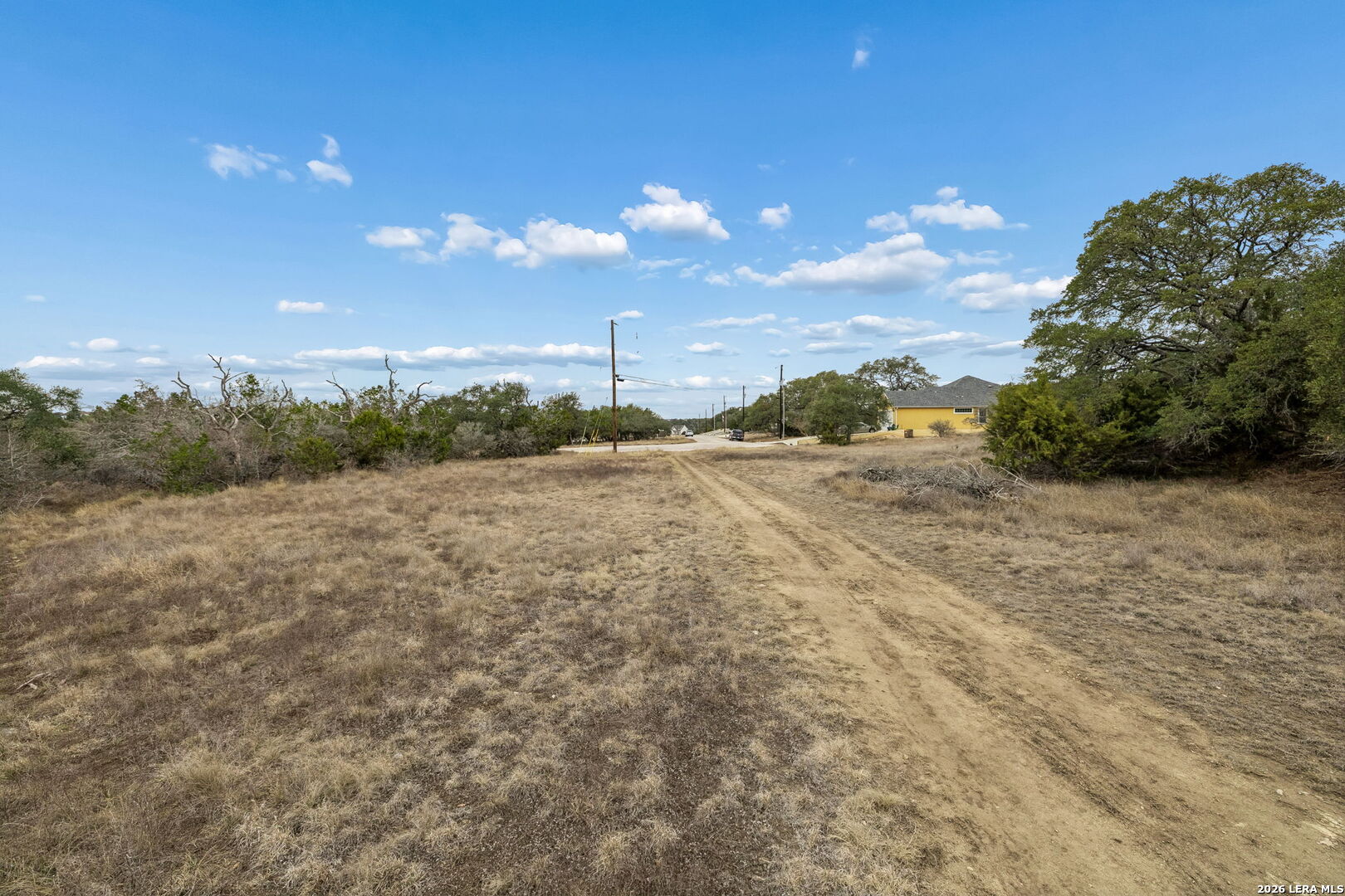 Open rural lot with dry grass and a dirt track leading toward a road and distant house.